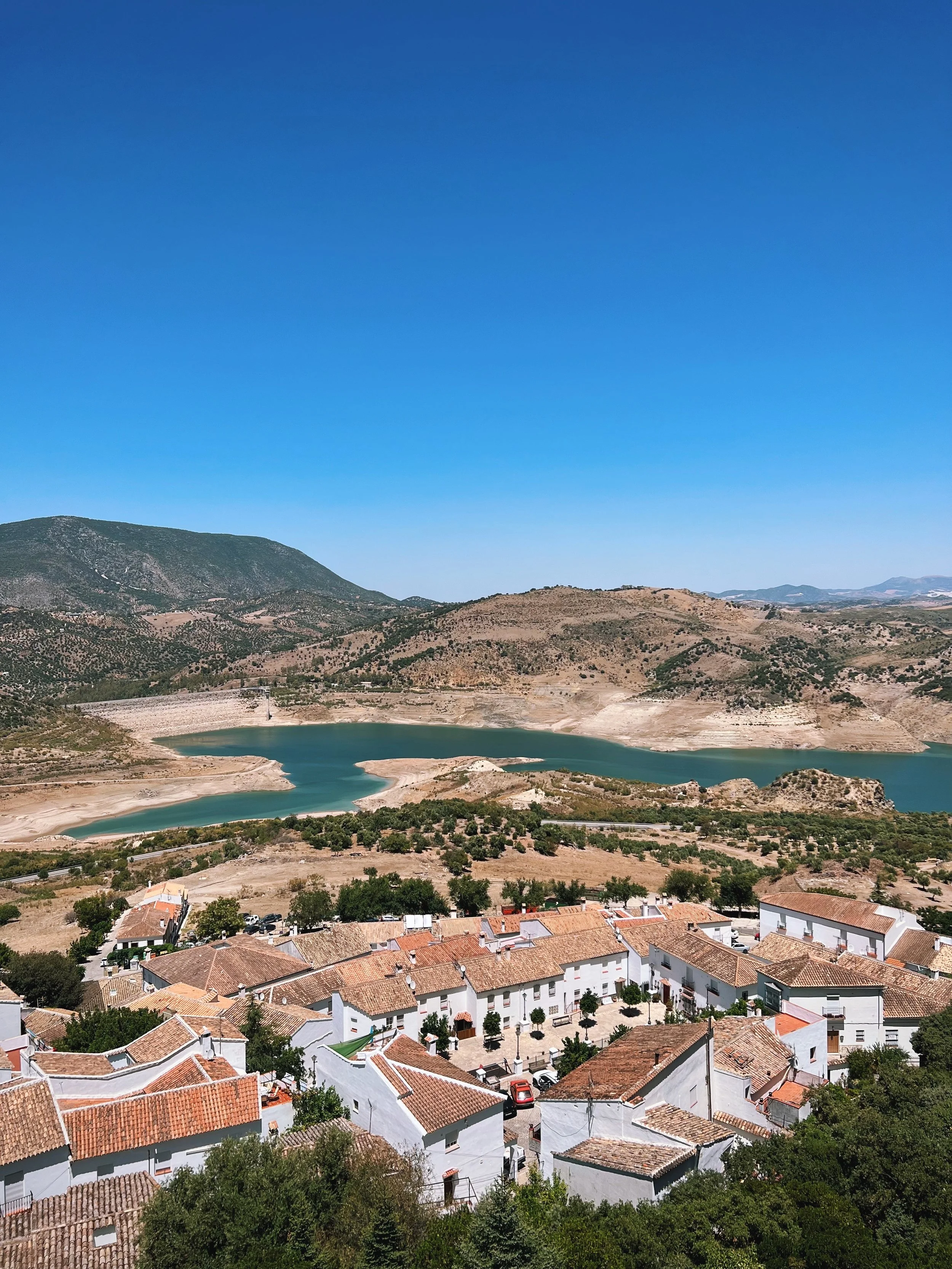 A scenic view of a town with white houses and terracotta roofs, situated near a large reservoir with surrounding hills and mountains under a clear blue sky.