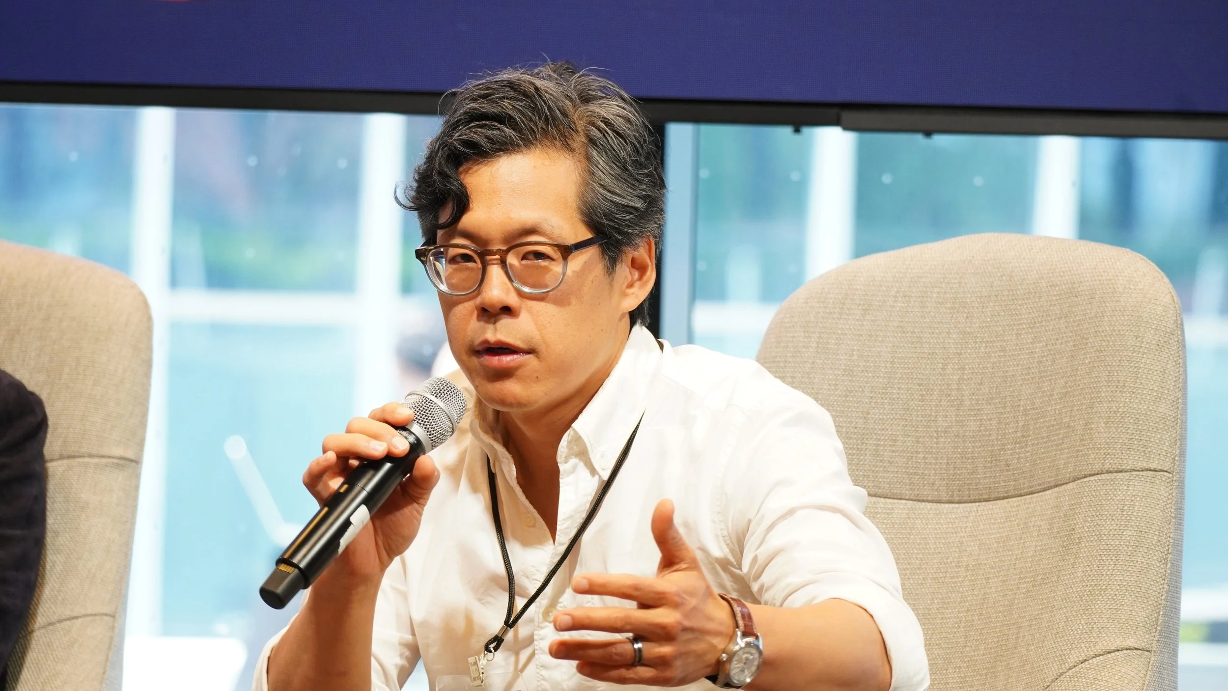 Man with glasses speaking into a microphone at a panel discussion or conference, seated on a beige chair, with a large window in the background.
