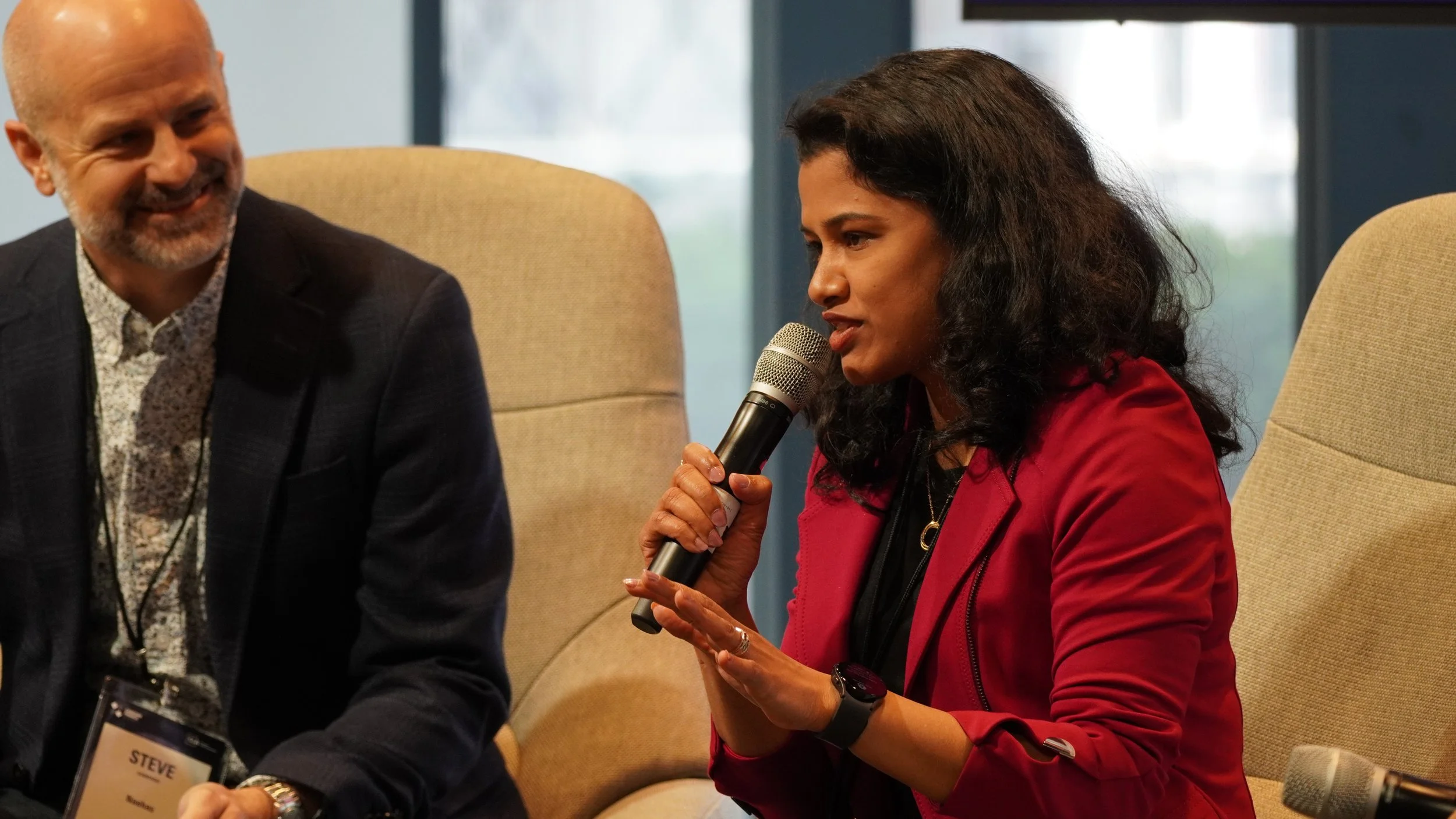 A woman in a red jacket holds a microphone and speaks during a panel discussion. A man in a suit sits beside her, smiling. They are seated in beige chairs, in a conference room with large windows in the background.