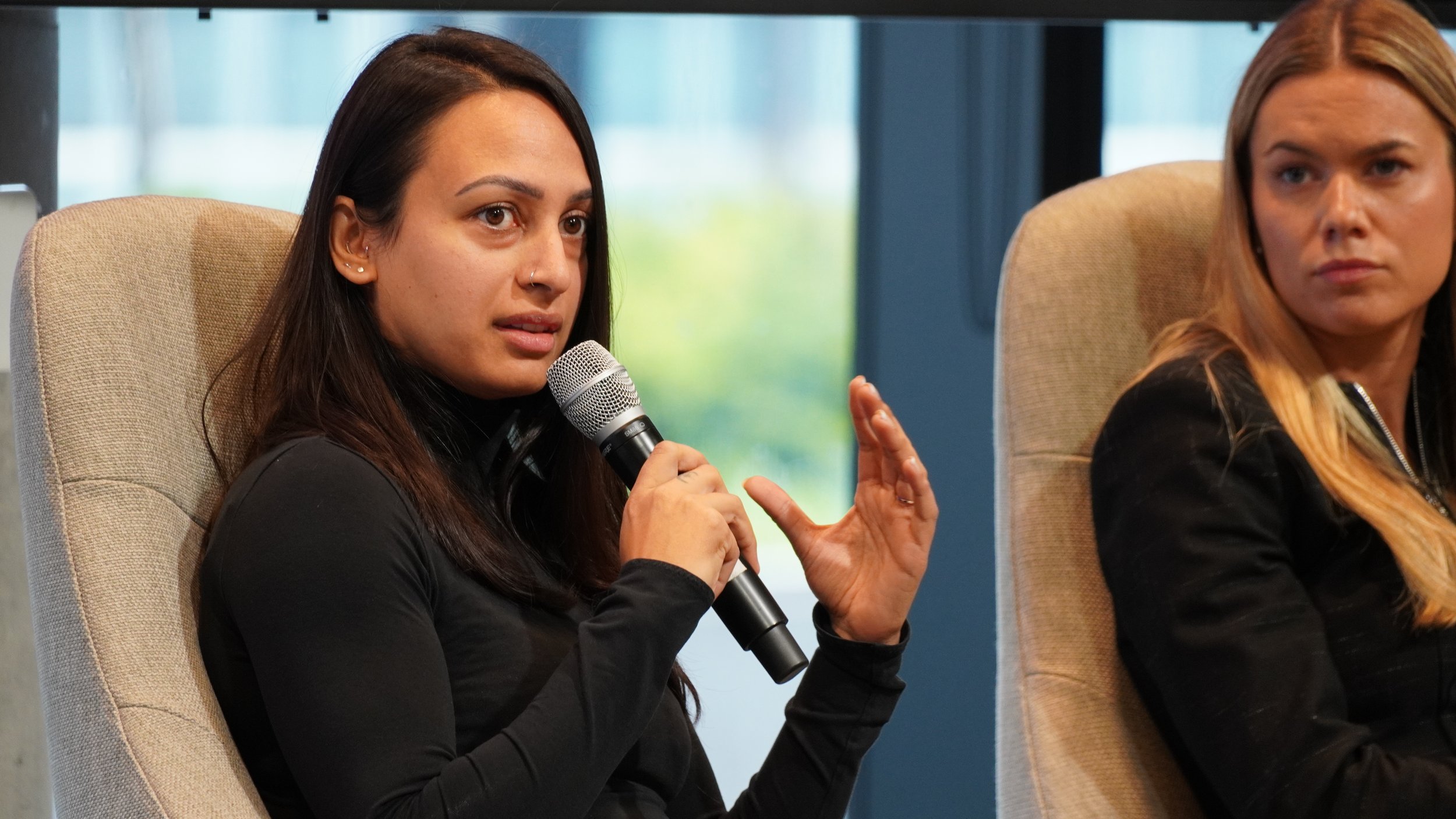 Woman with long dark hair and a nose ring speaking into a microphone during a panel or discussion, sitting next to another woman with long blonde hair in a black jacket.