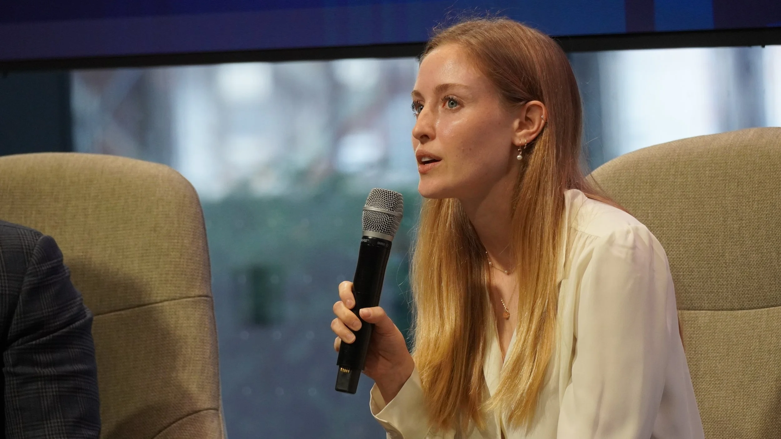 A woman with long red hair, wearing a white blouse and pearl earrings, is speaking into a microphone during a panel discussion or interview. She is seated on a beige chair with a blurred outdoor background visible through a window.