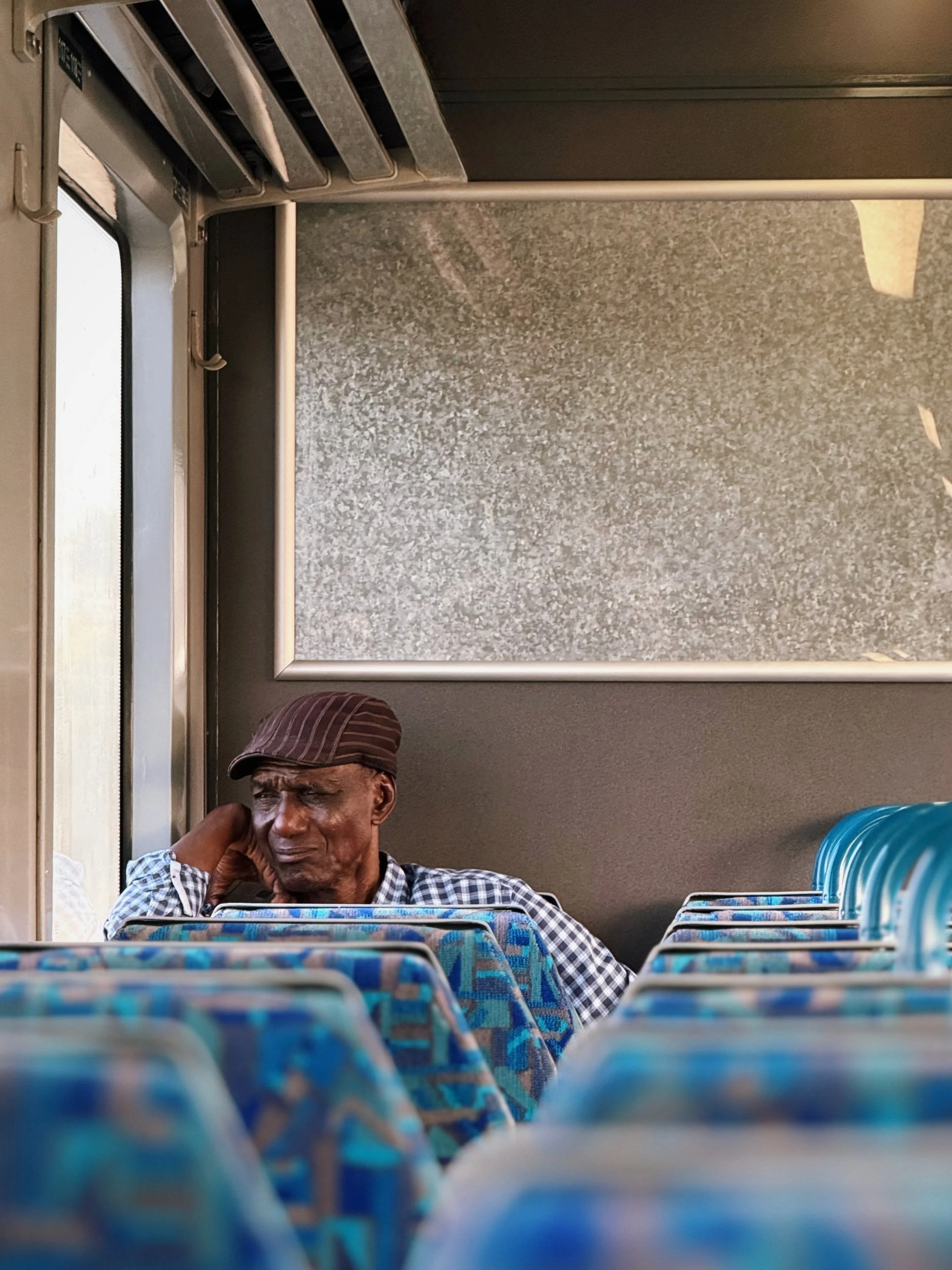 An elderly man sitting alone on a bus, resting his head on his hand with a thoughtful or tired expression, wearing a striped cap and checkered shirt.