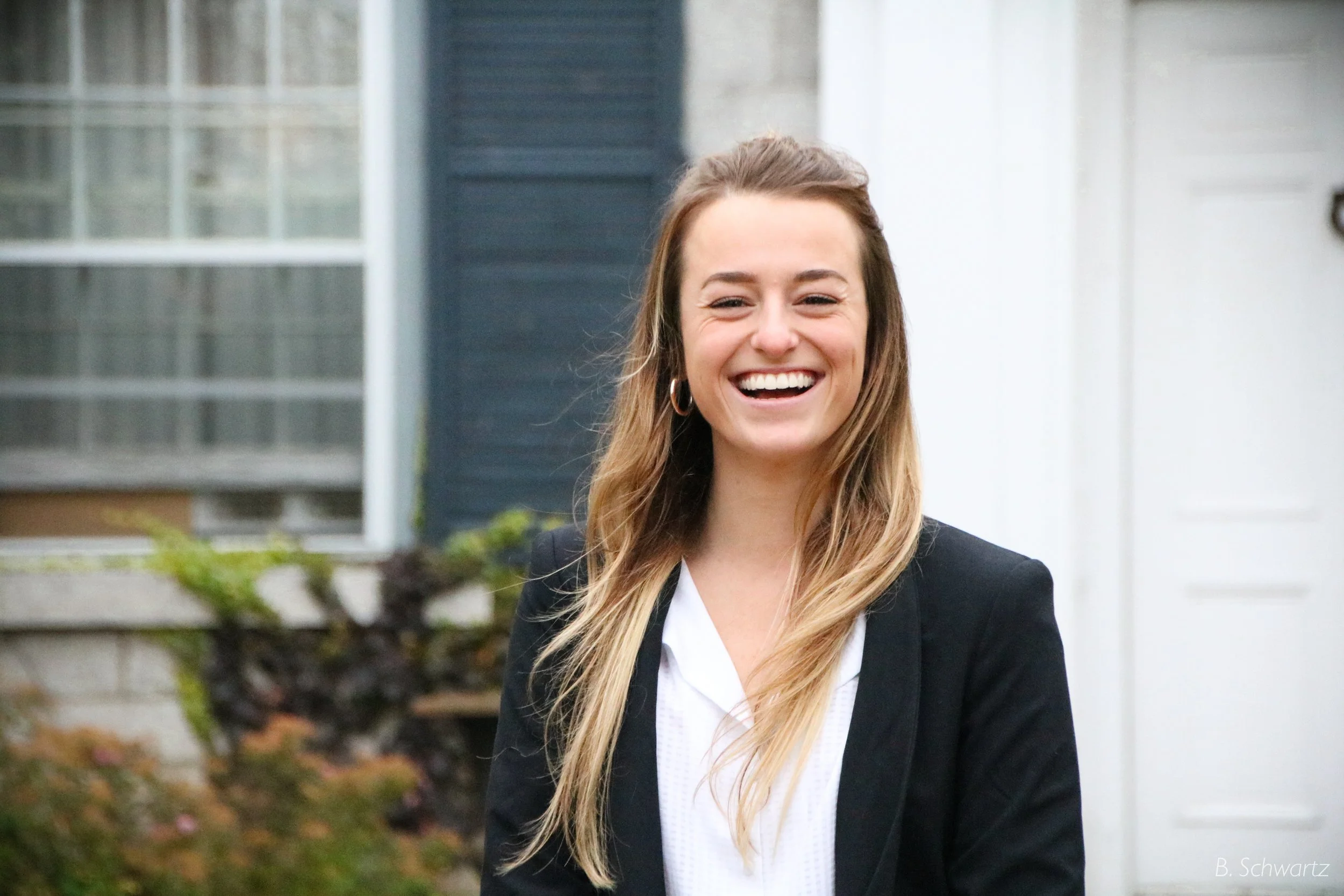 A young woman in business attire is smiling outdoors in front of a house with a window and plants.