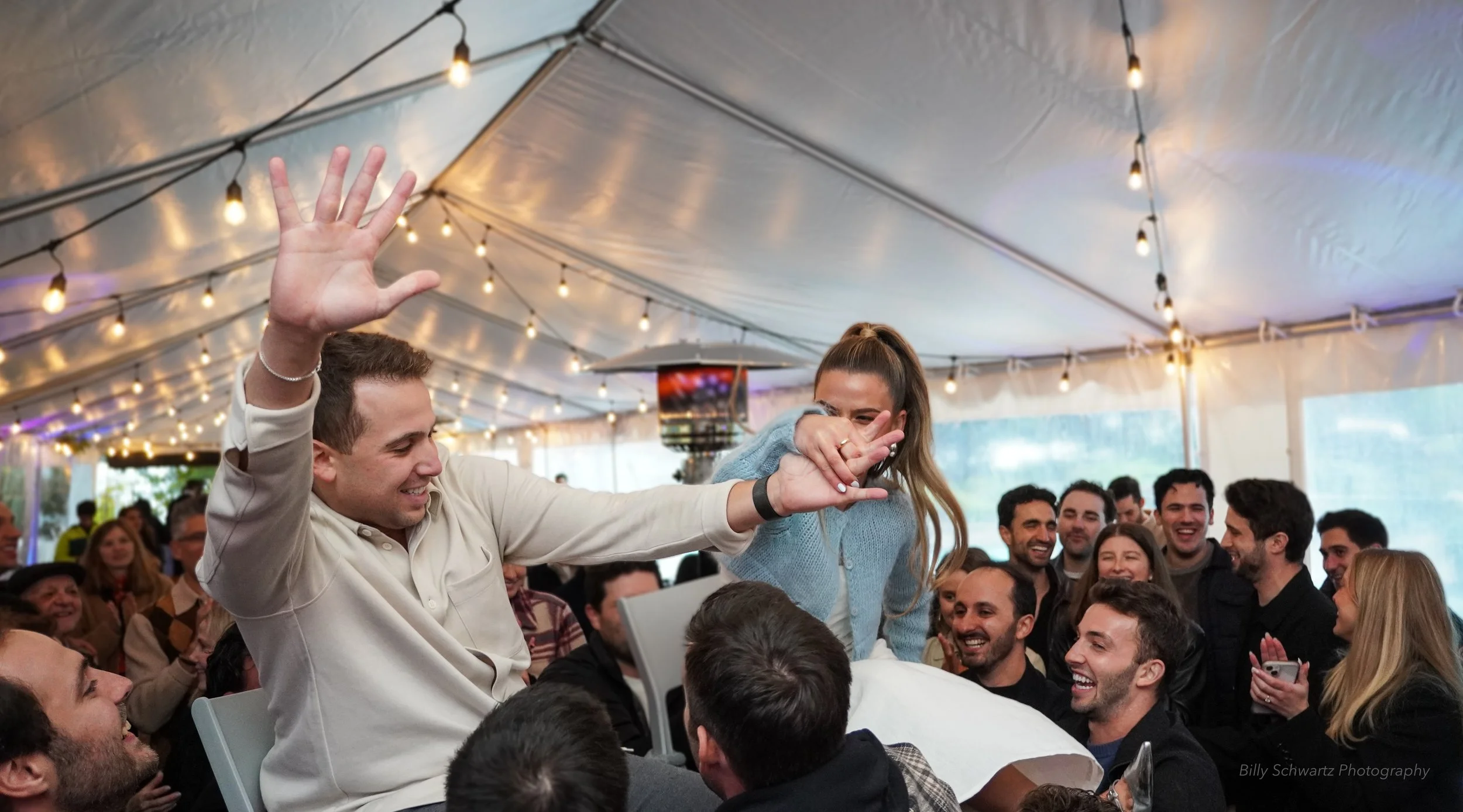 People celebrating at a festive event under a tent, with string lights overhead. A man and woman are in the center, standing and smiling, surrounded by a crowd of laughing and happy individuals.