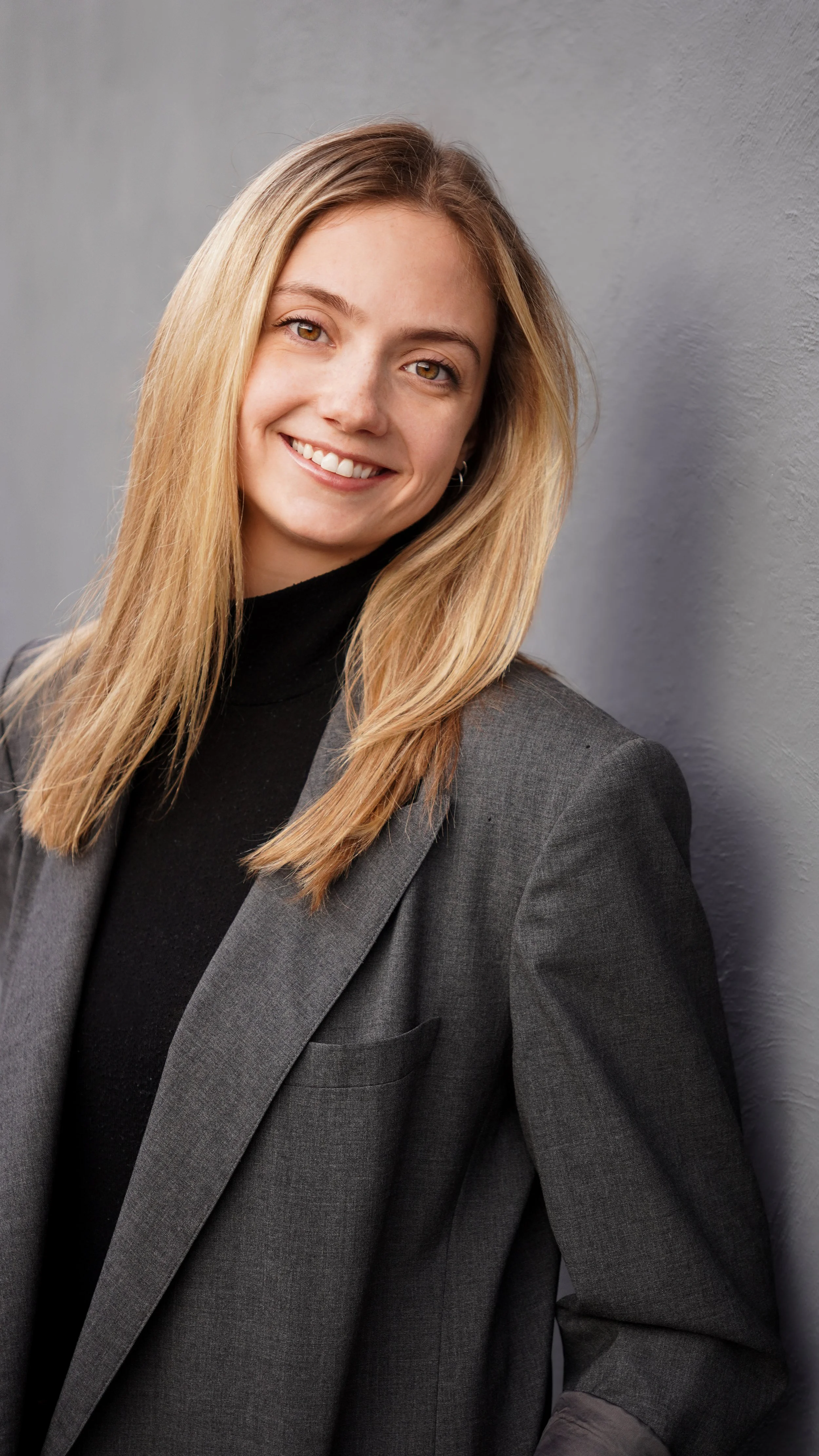 A young woman with long blonde hair, wearing a dark blazer and black turtleneck, smiling and leaning against a gray wall.