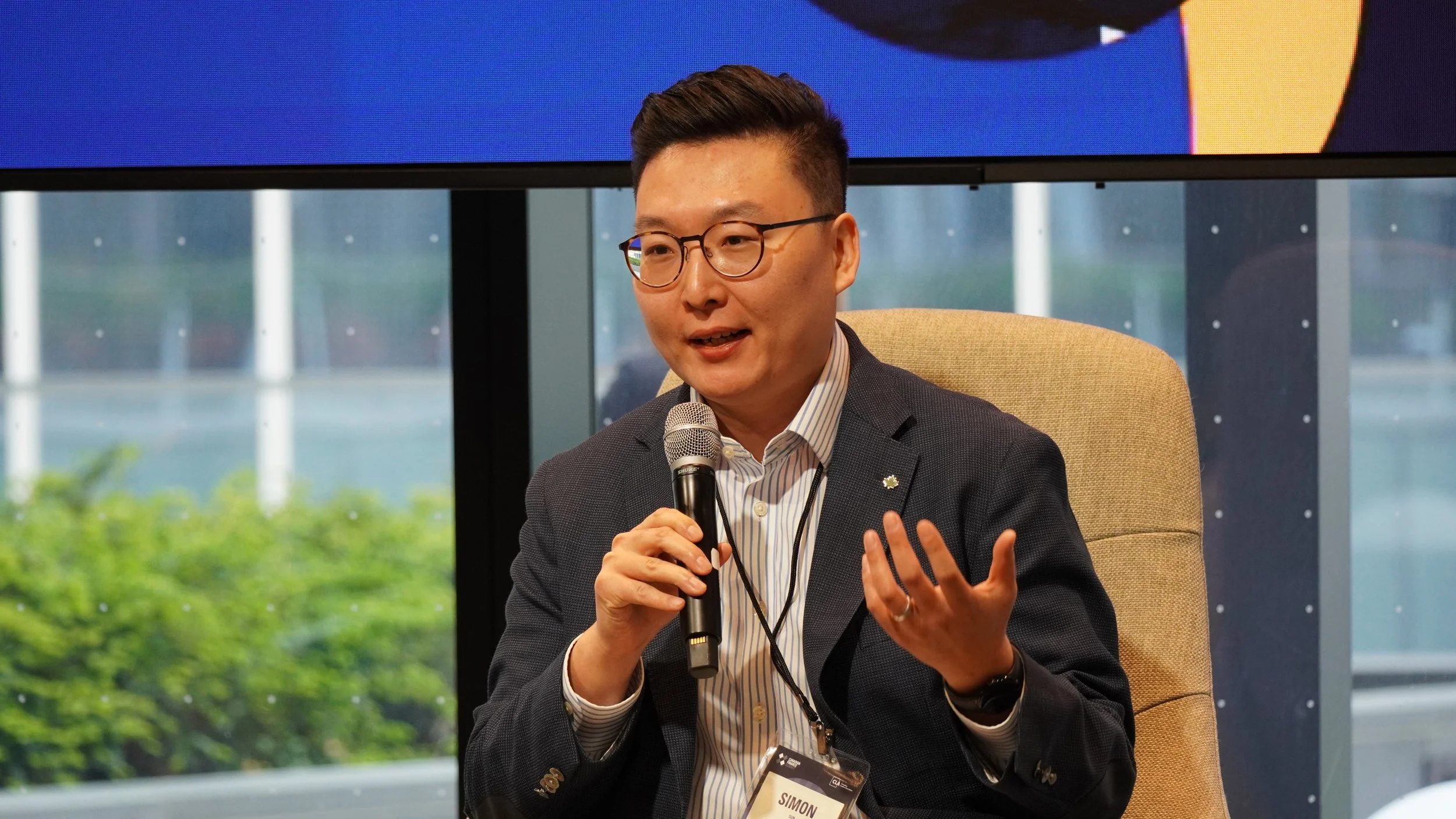 Man in glasses holding a microphone and speaking at an indoor event, wearing a dark blazer, striped shirt, and a name tag that says 'Simon'.
