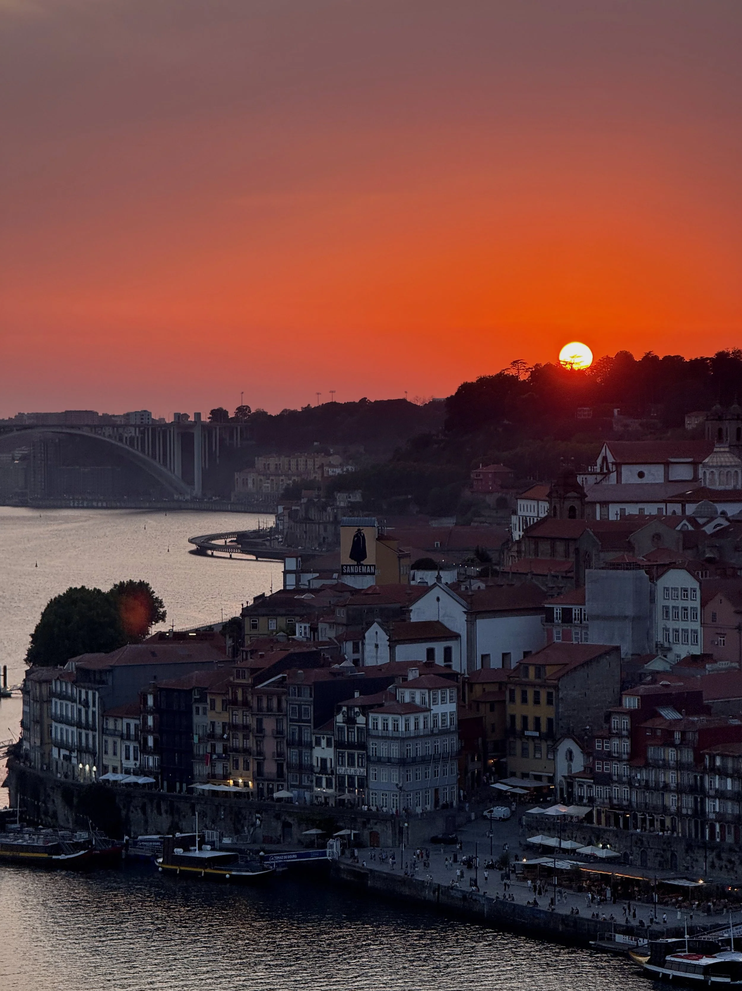 Sunset over a riverside city with historic buildings, a bridge, and a hillside with trees.