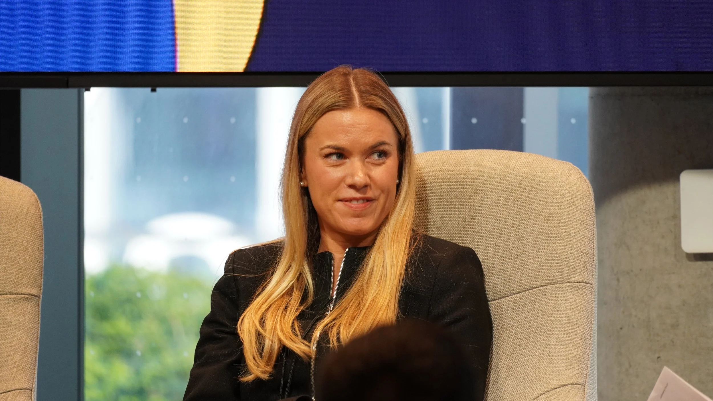A woman with long blonde hair seated in a beige chair, wearing a black jacket, looking to her right with a slight smile. There is a window behind her showing a rainy outdoor scene and a large screen above her with colorful graphics.