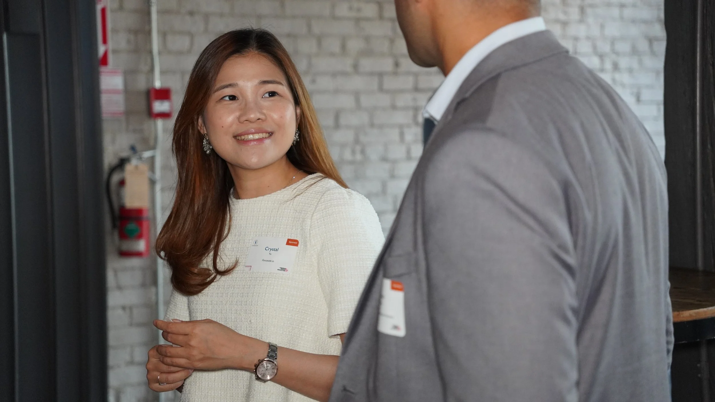 A young woman with long brown hair wearing a white blouse, earrings, and a watch is smiling while talking to a man in a gray suit. They are indoors with a brick wall and fire safety equipment in the background.