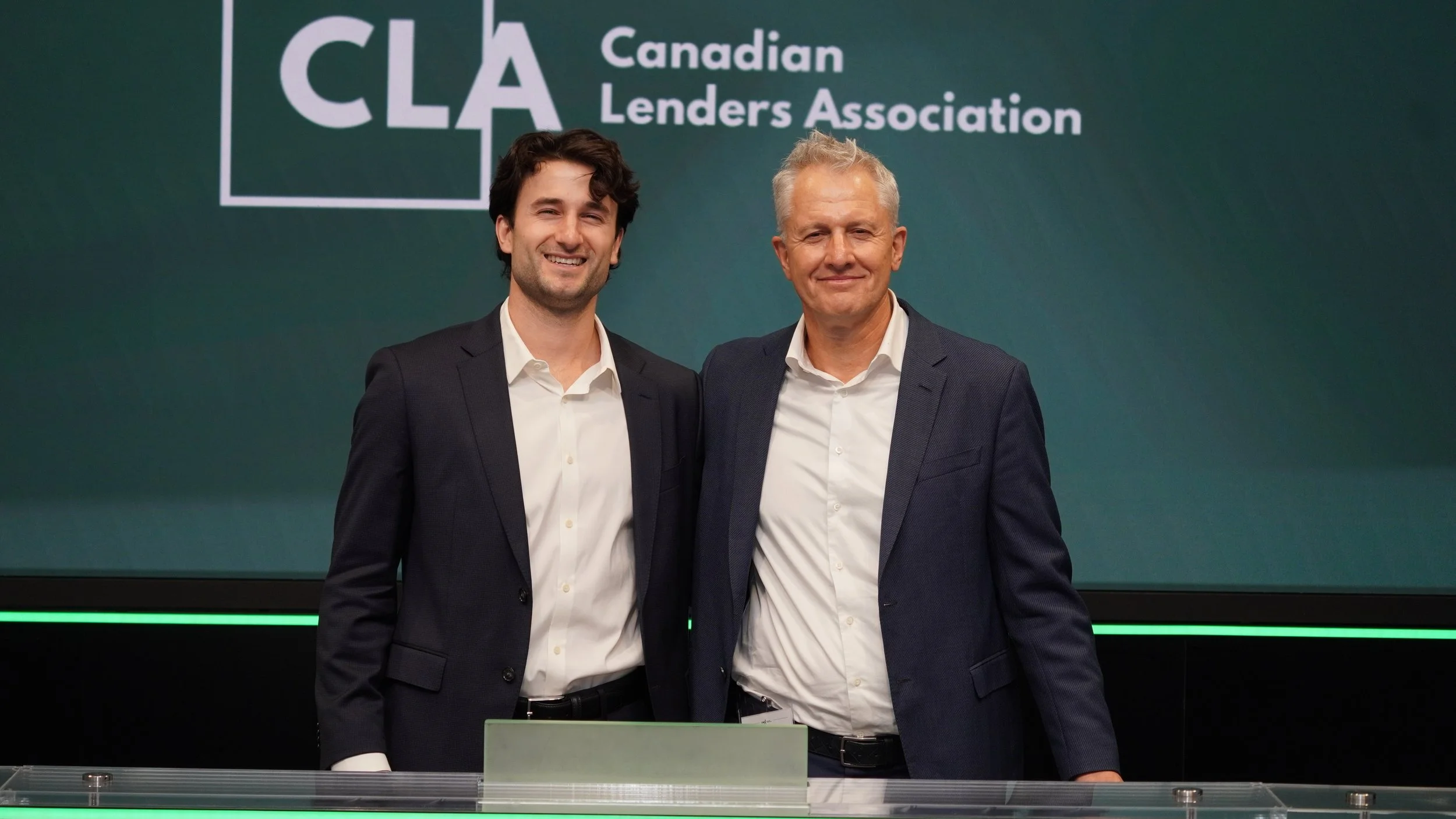 Two men in suits standing together smiling in front of a backdrop with the text 'CLA Canadian Lenders Association'