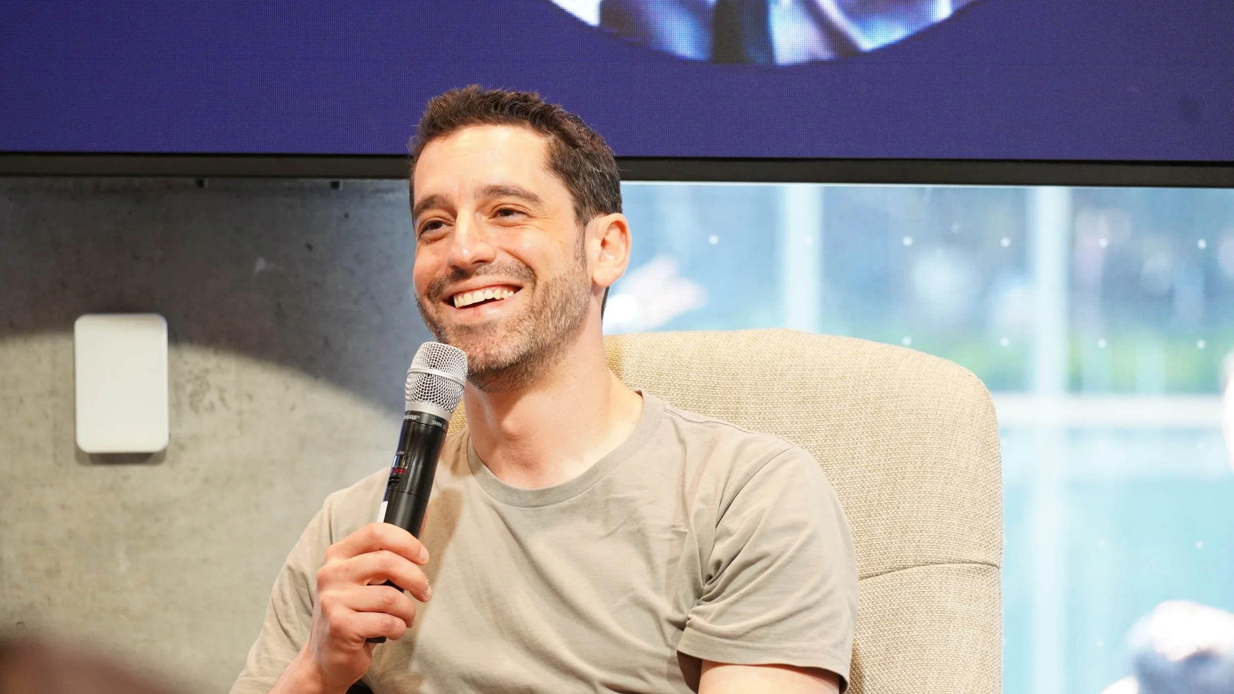 A smiling man with dark hair and a beard, wearing a light gray t-shirt, sitting on a beige chair, holding a microphone, during a speaking event or interview, with a blurred cityscape visible through large windows in the background.