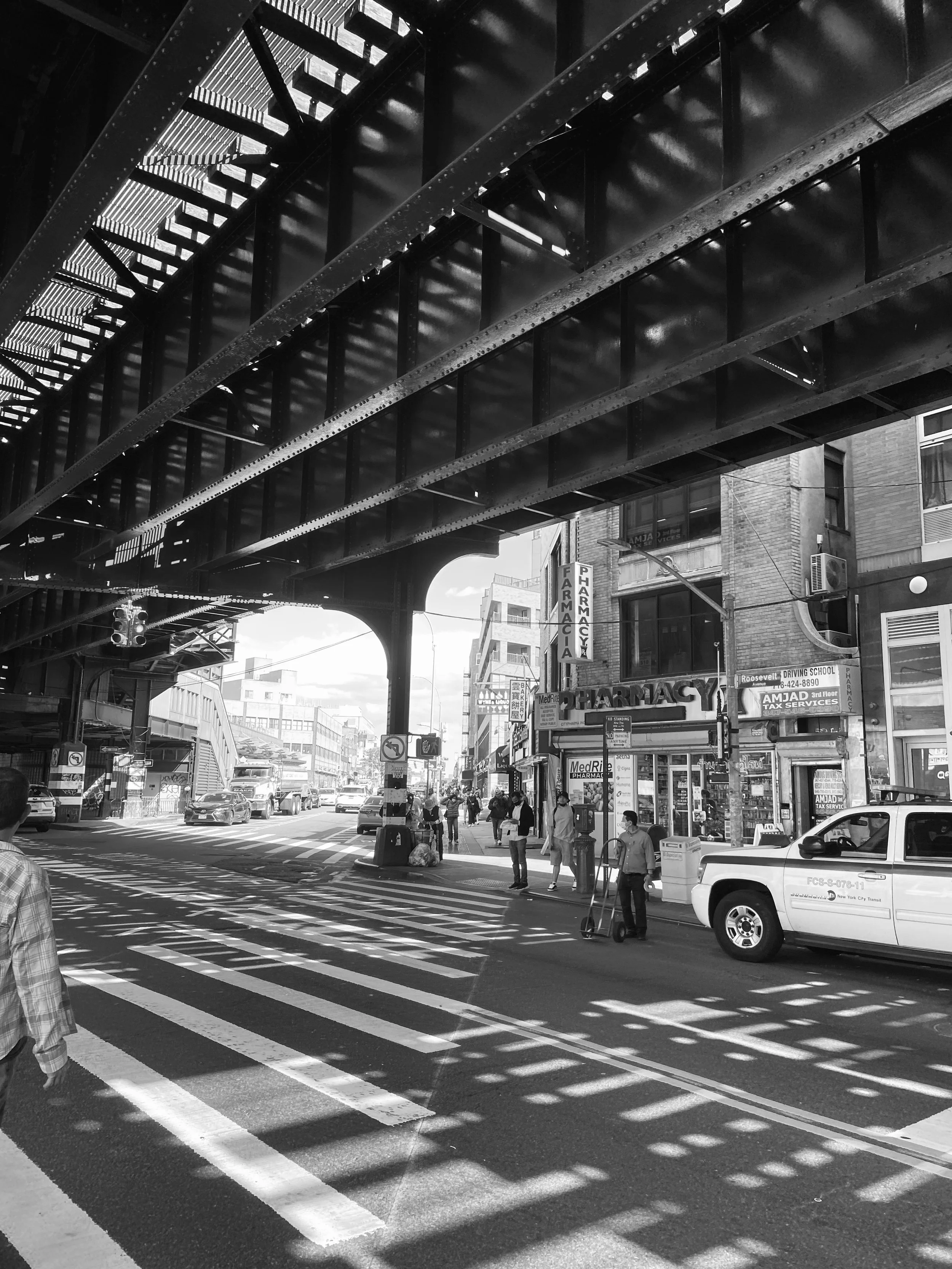 Street scene in an urban area showing a crosswalk, a pharmacy, pedestrians, and vehicles under an elevated train track.