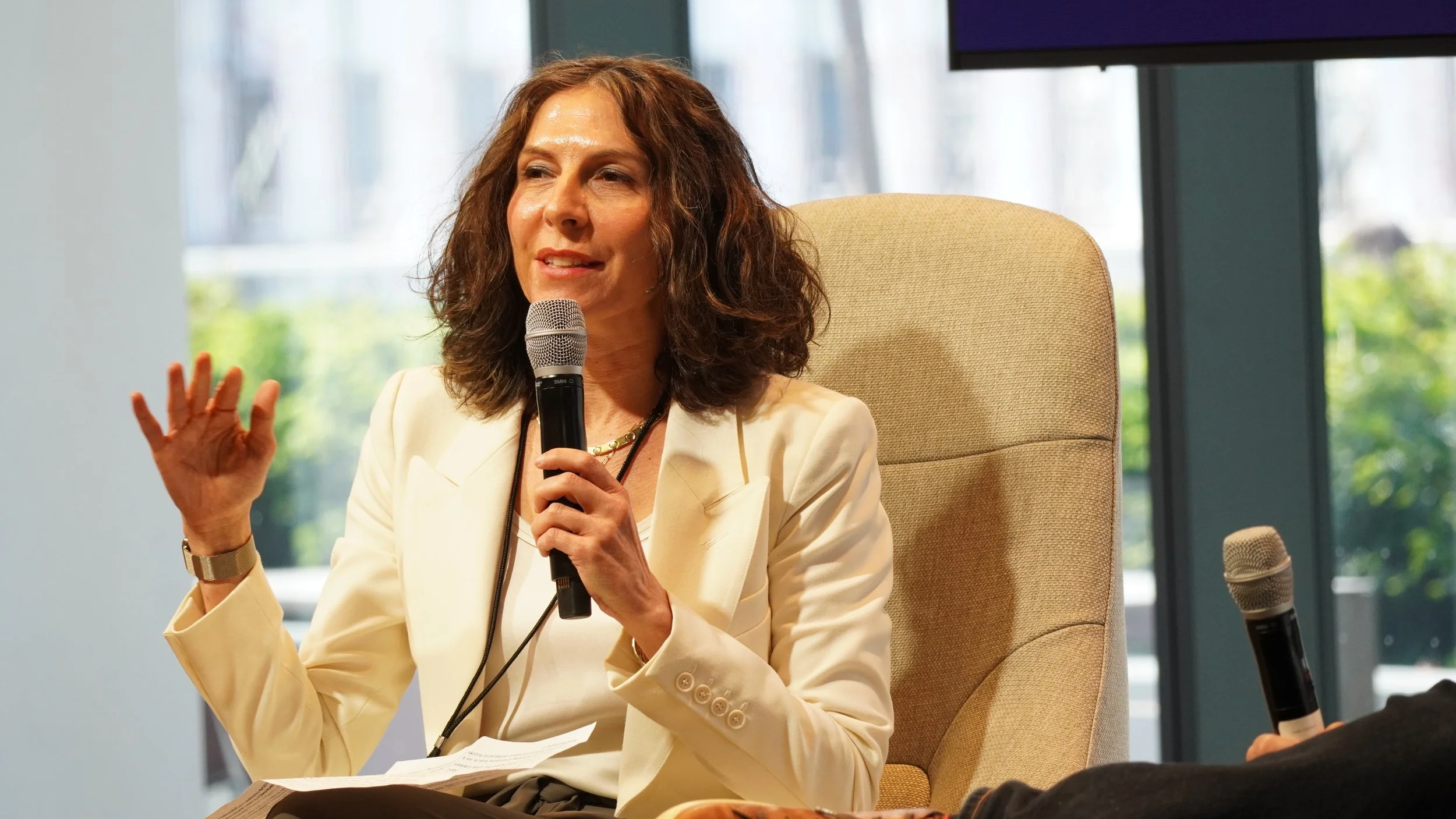 Woman with curly hair wearing a cream blazer, speaking into a microphone during a panel or discussion, sitting on a beige chair with a window in the background showing trees.
