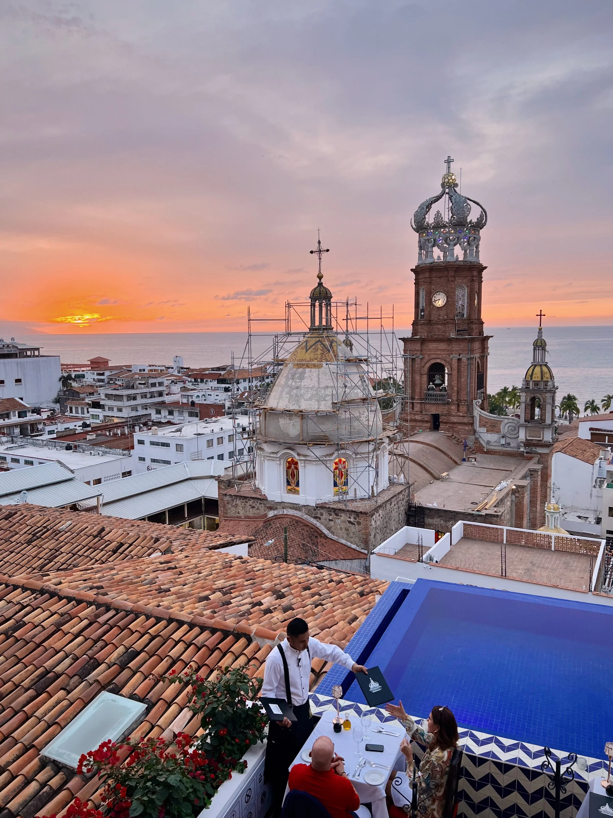 People enjoying dinner at a rooftop restaurant overlooking a city with churches and the ocean during sunset in Mexico.