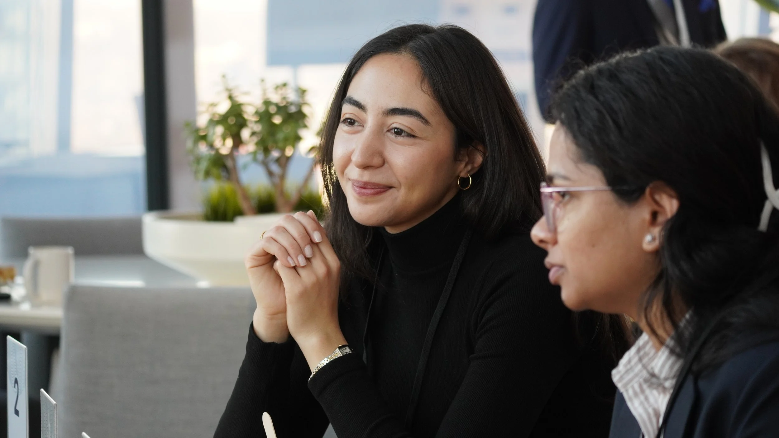 Two women sitting at a table in a meeting room, one smiling with hands folded, the other wearing glasses and looking to the side, with a potted plant and large windows in the background.