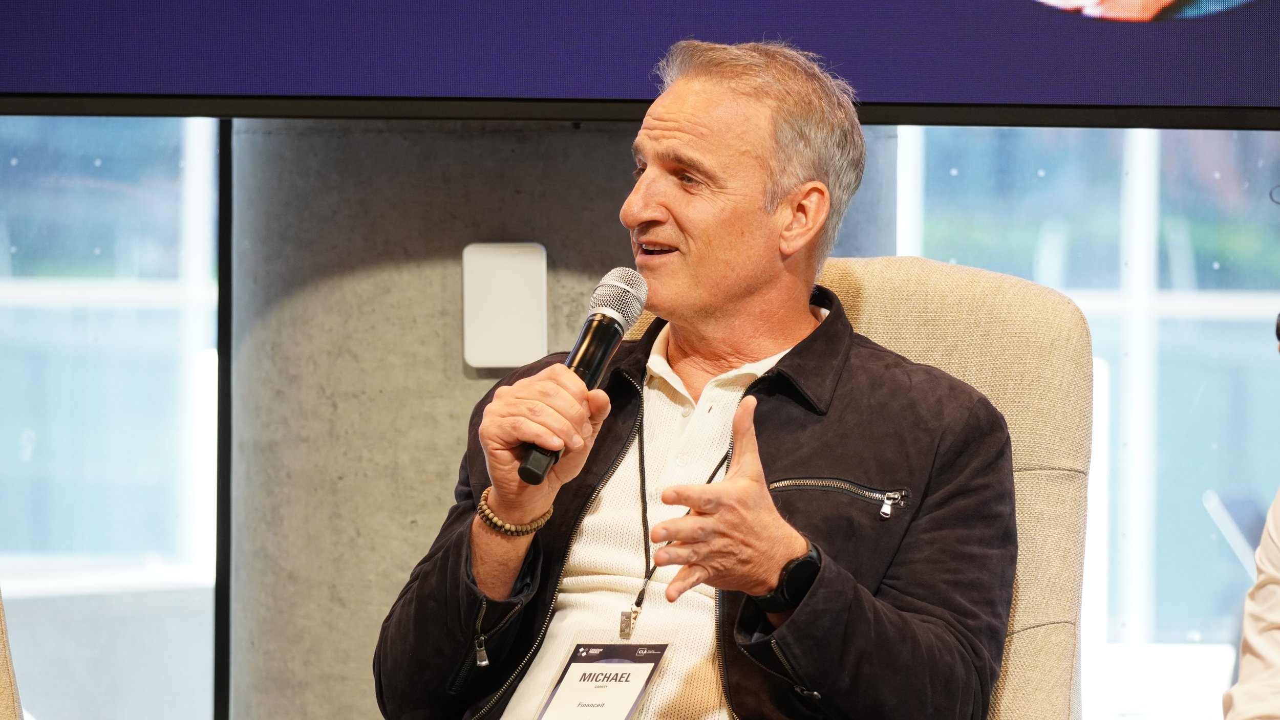 A man with gray hair, wearing a black jacket, white polo shirt, and a wristwatch, is sitting on a beige chair, speaking into a microphone, during an indoor event.