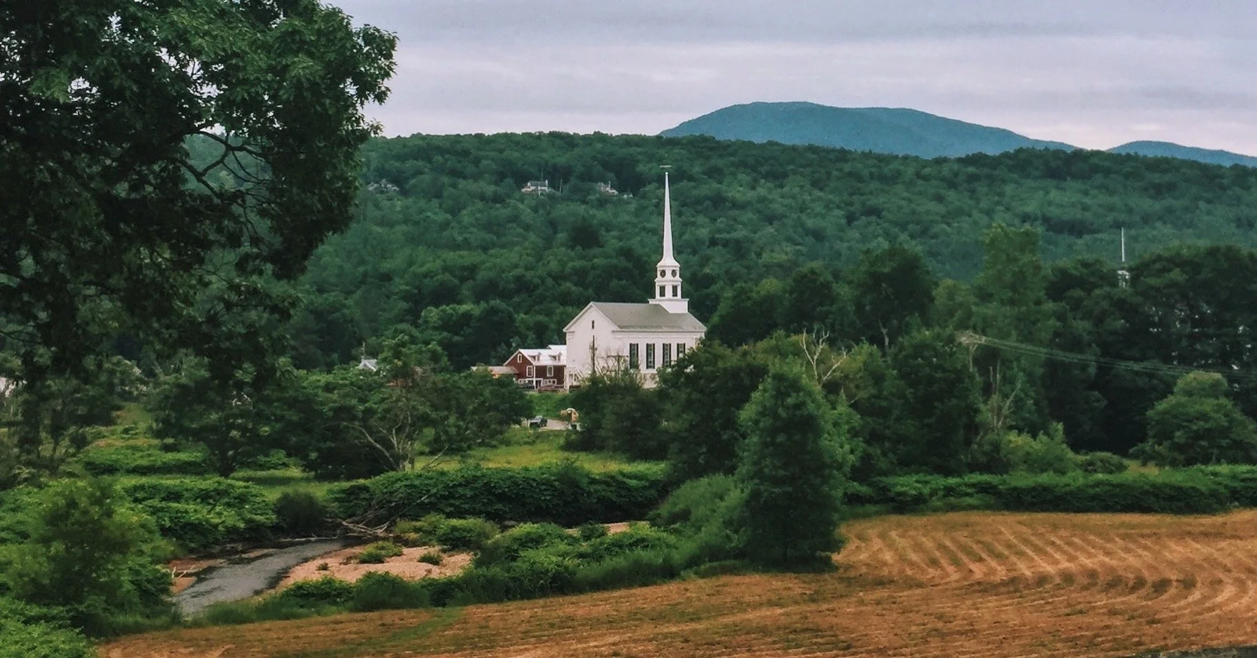 A white church with a tall steeple situated amidst lush green trees and rolling hills under an overcast sky.