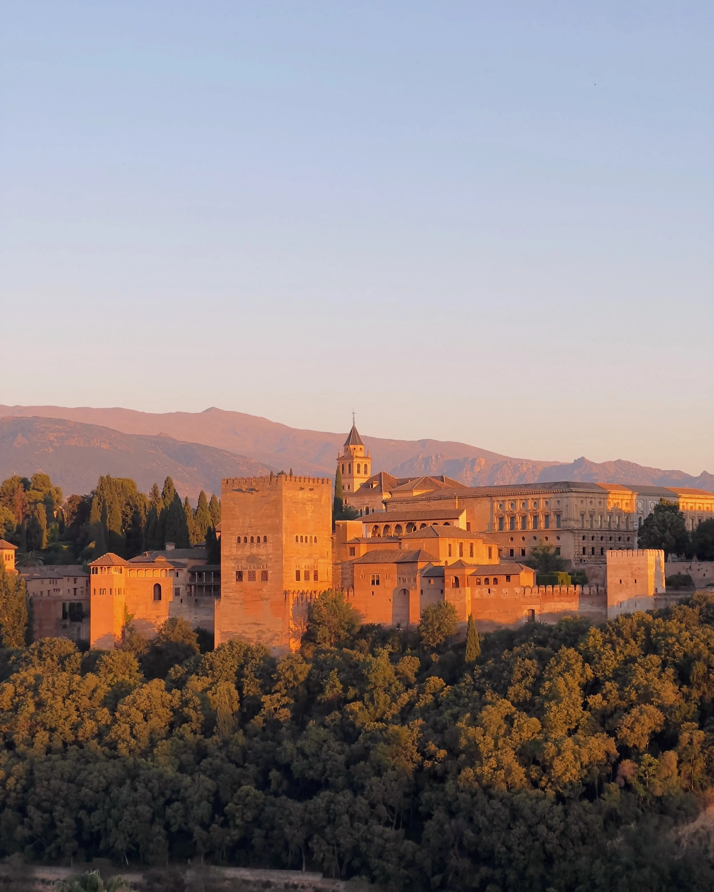 View of a historic castle with towers and walls, surrounded by trees and hills at sunset.