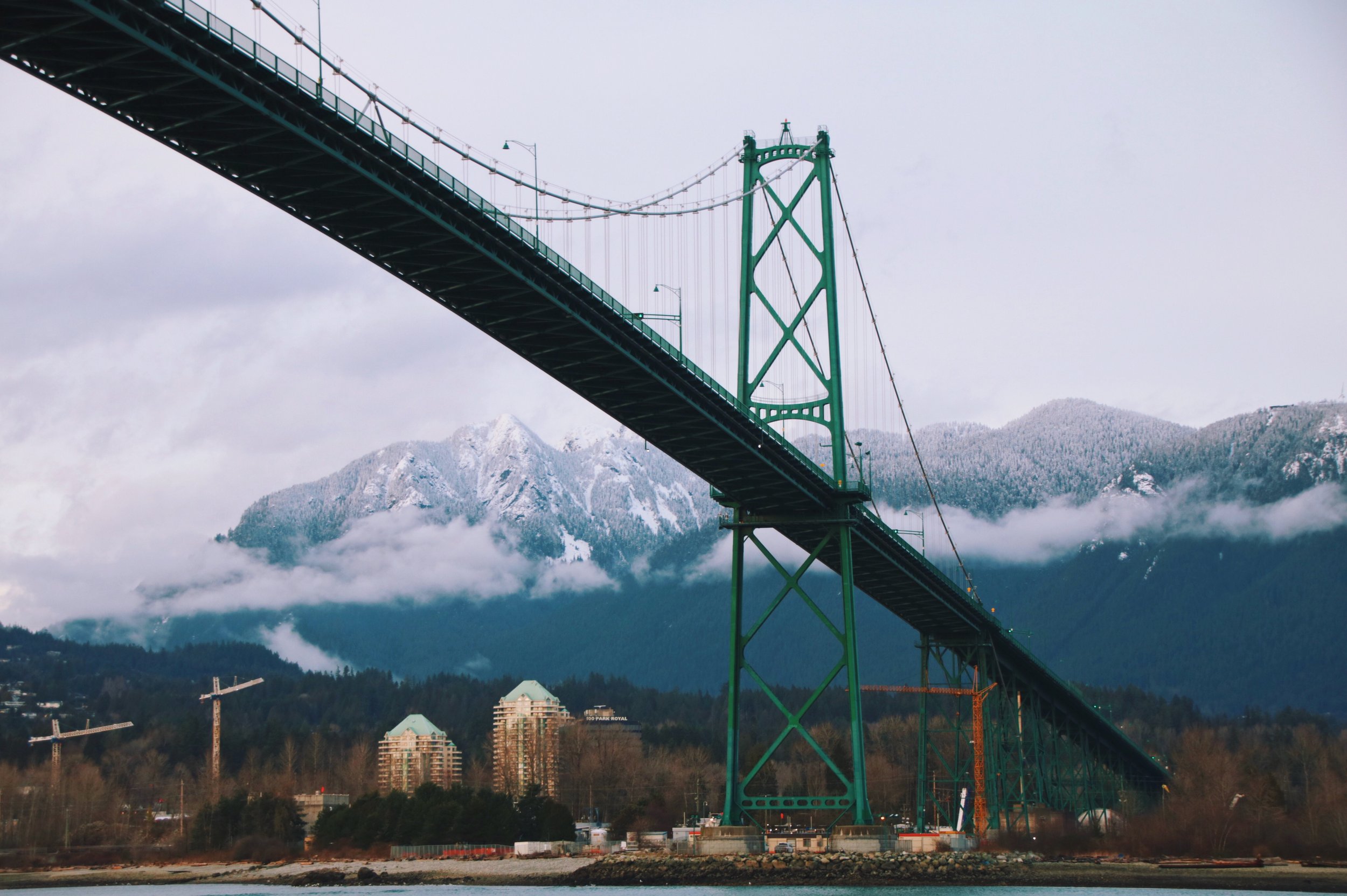 A large suspension bridge with green towers extending over a body of water, mountain peaks with snow and clouds in the background, and buildings and trees in the foreground.