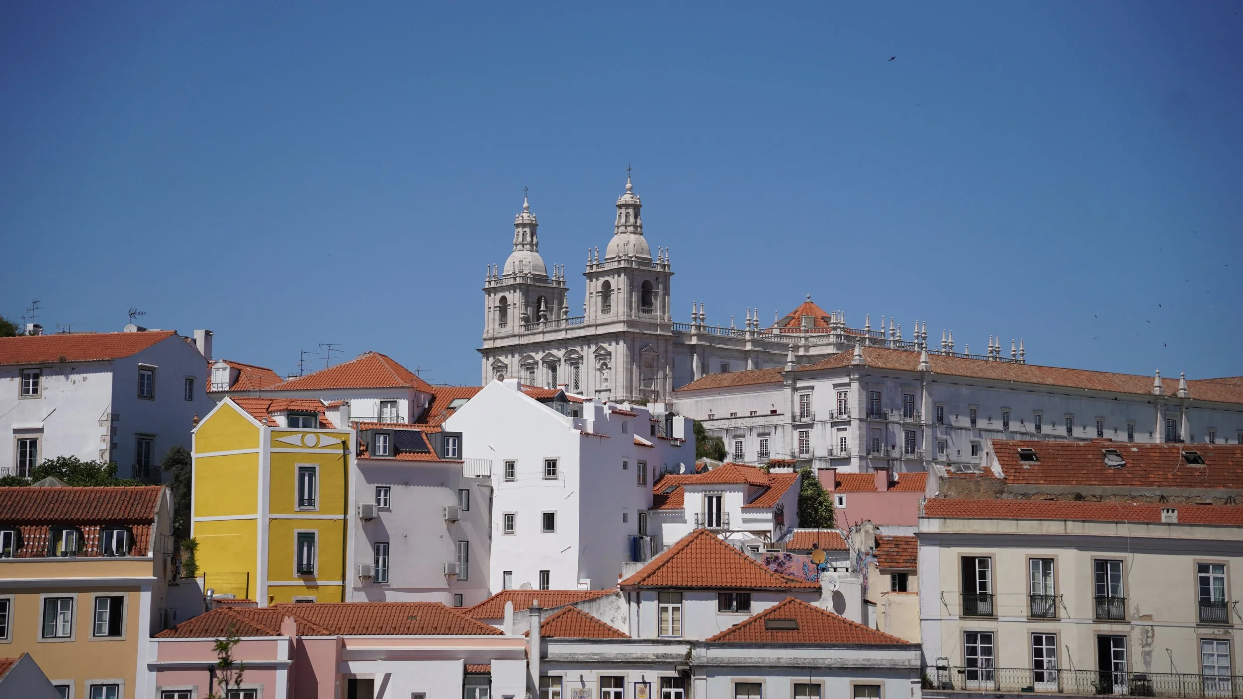 Cityscape of Lisbon, Portugal, featuring colorful buildings with red-tiled roofs and a historic cathedral with twin towers in the background, against a clear blue sky.