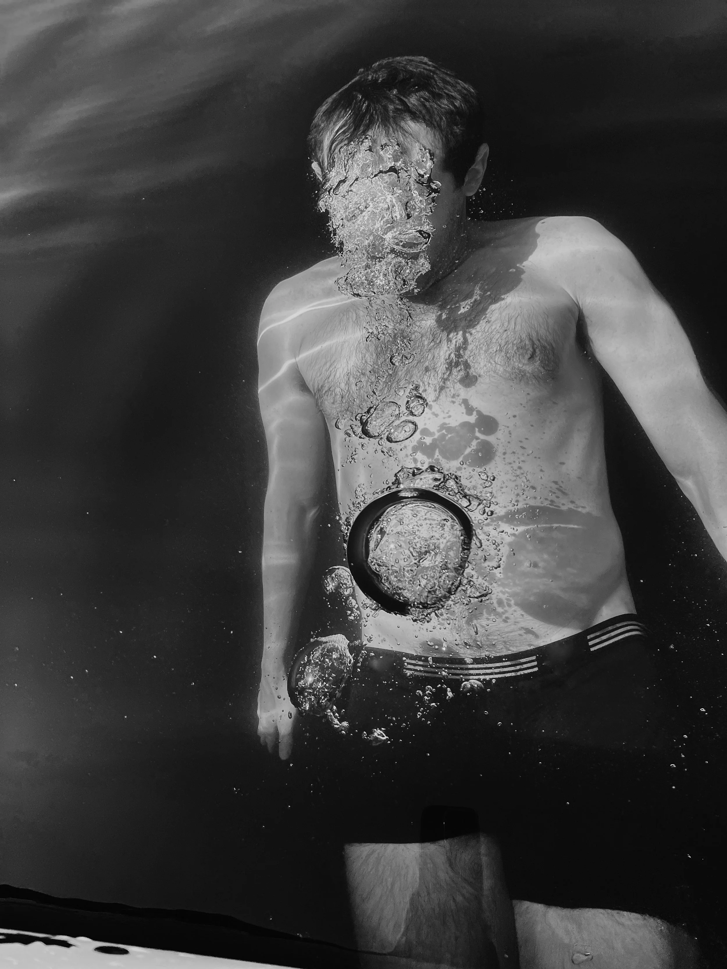 A man floating in water with his face underwater, surrounded by bubbles, wearing black swim trunks.