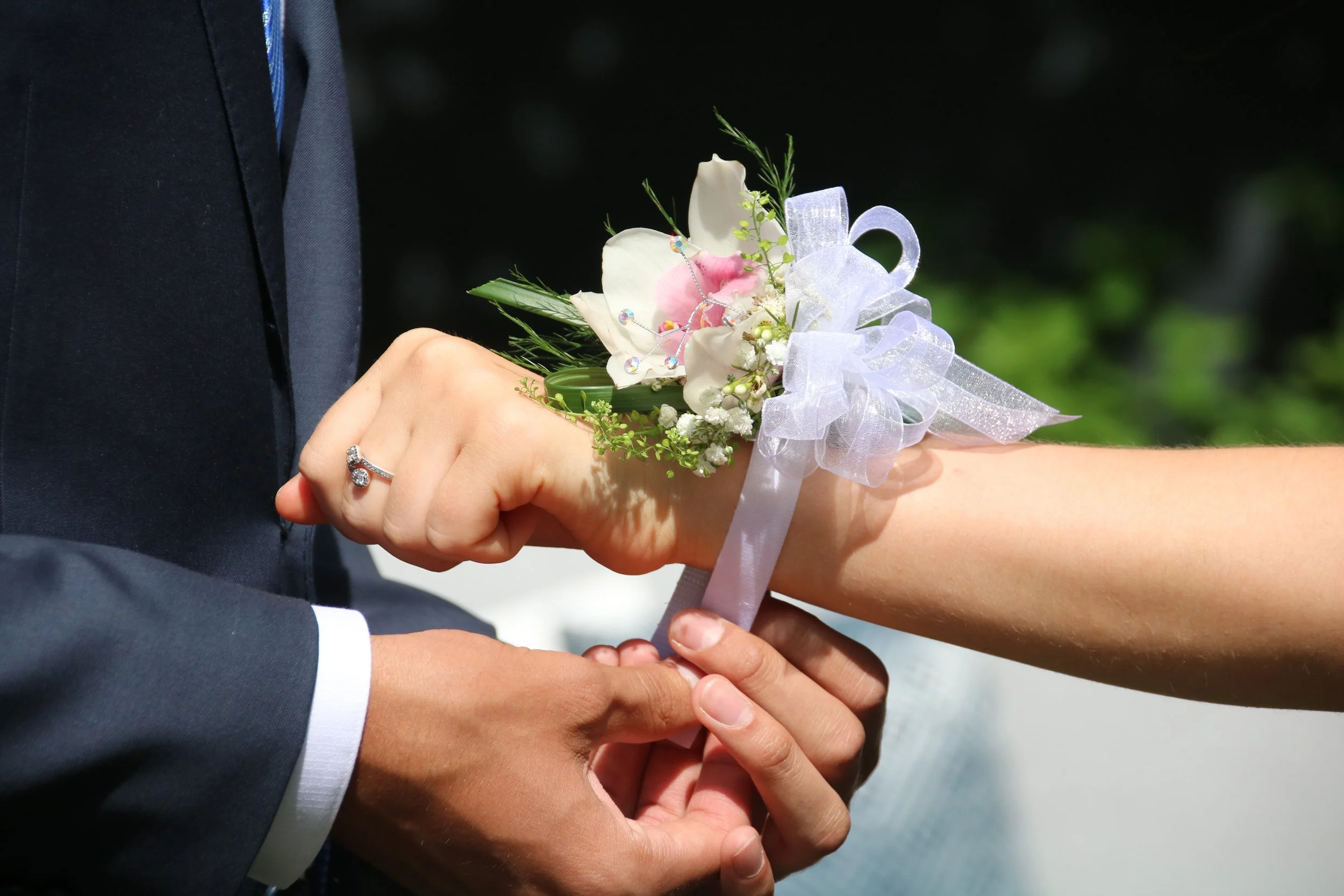 Close-up of a couple holding hands during a wedding, with the woman giving the man a floral boutonniere. The woman wears a wedding ring, and the boutonniere has white flowers, greenery, and a white ribbon.