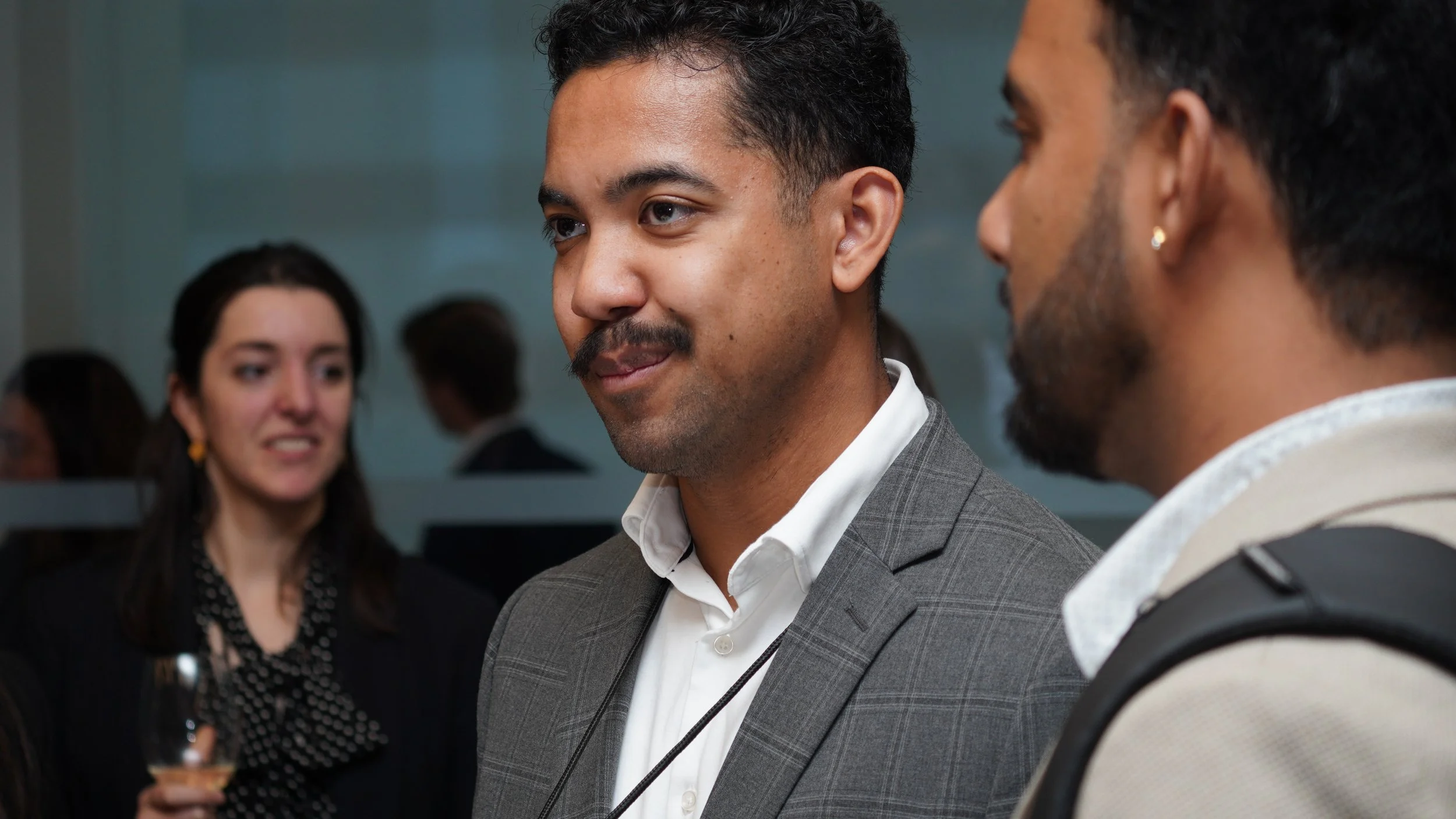 Two men engaged in conversation at a professional event, with a woman in the background holding a glass of wine.