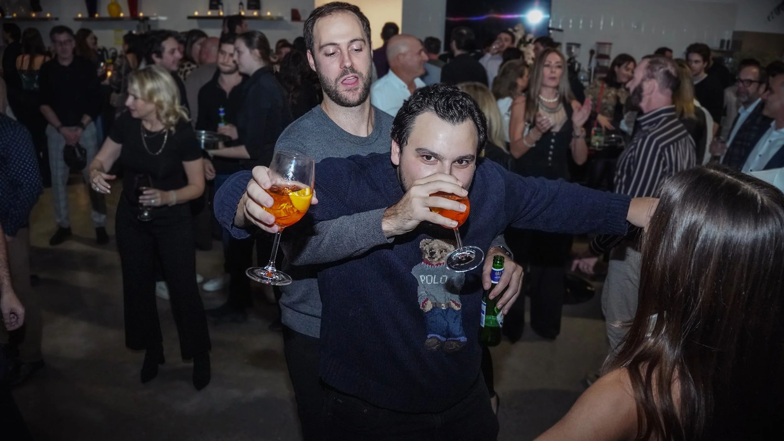 People at a social gathering, two men in the foreground are drinking cocktails, one man is holding a glass with an orange slice and the other is drinking from a glass, surrounded by other people socializing.