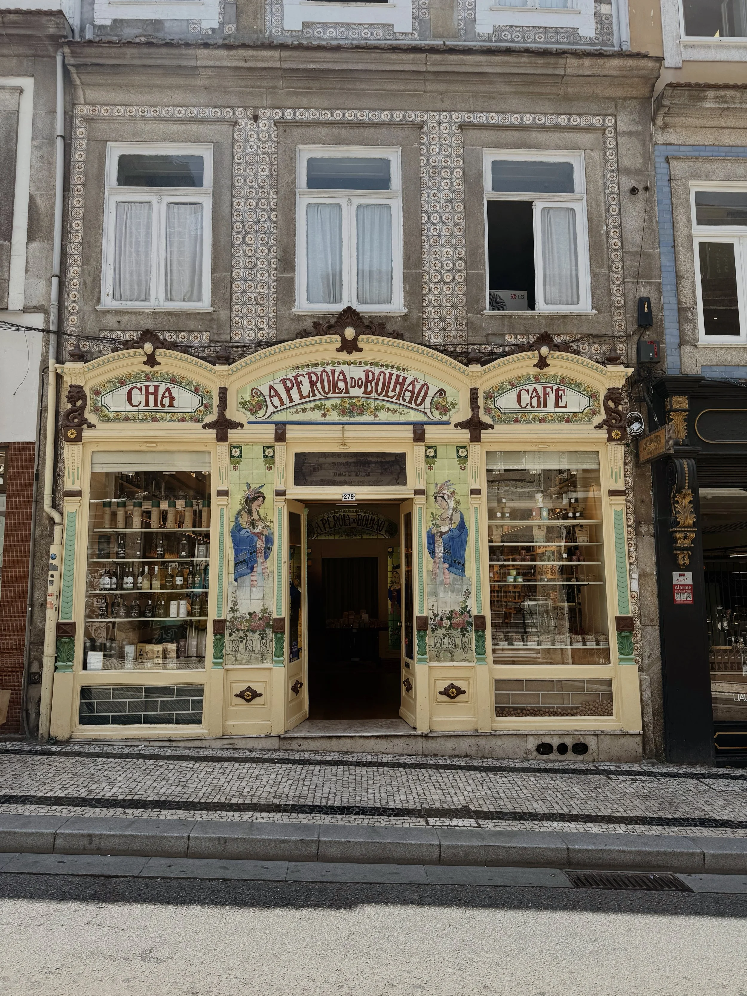 The storefront of a shop named "A Perola do Bolhao" that sells tea and coffee. The shop has an ornate, vintage design with painted images of women, and displays bottles and jars in the window.