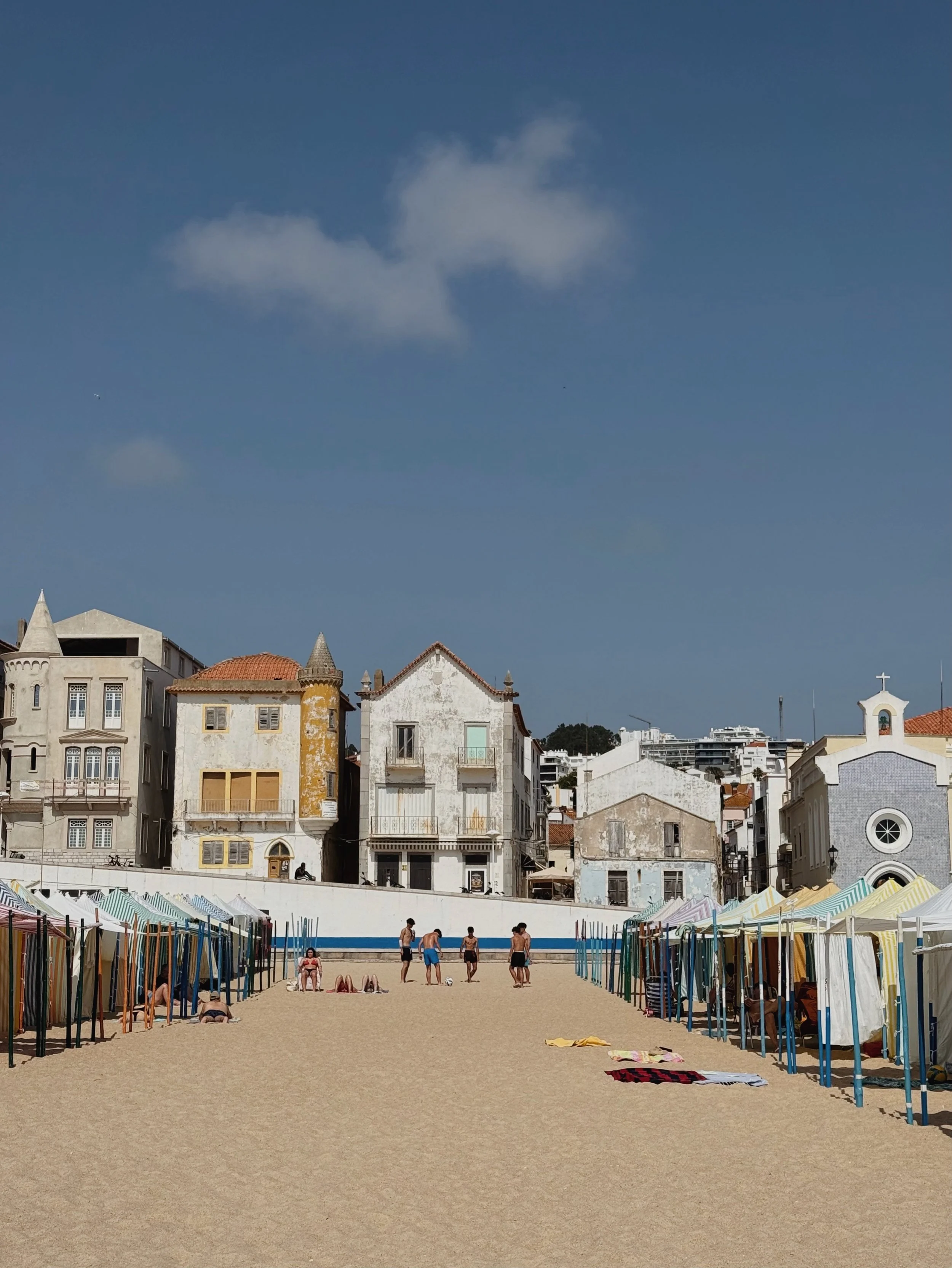Beach with tents and people, backlit by old buildings and a blue sky with a few clouds.