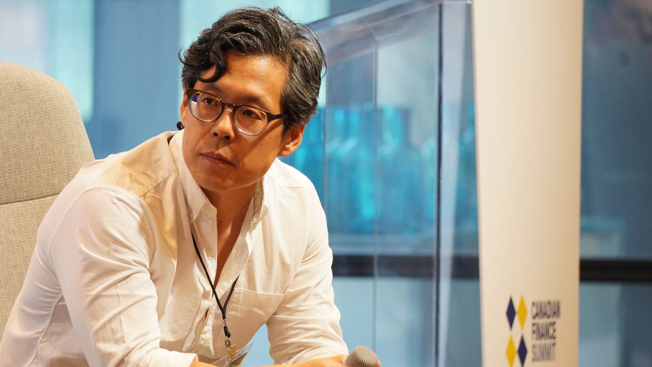 Man with glasses sitting on a beige chair, holding a microphone, at an indoor event with a sign that reads "Sustainable Finance Summit" in the background.