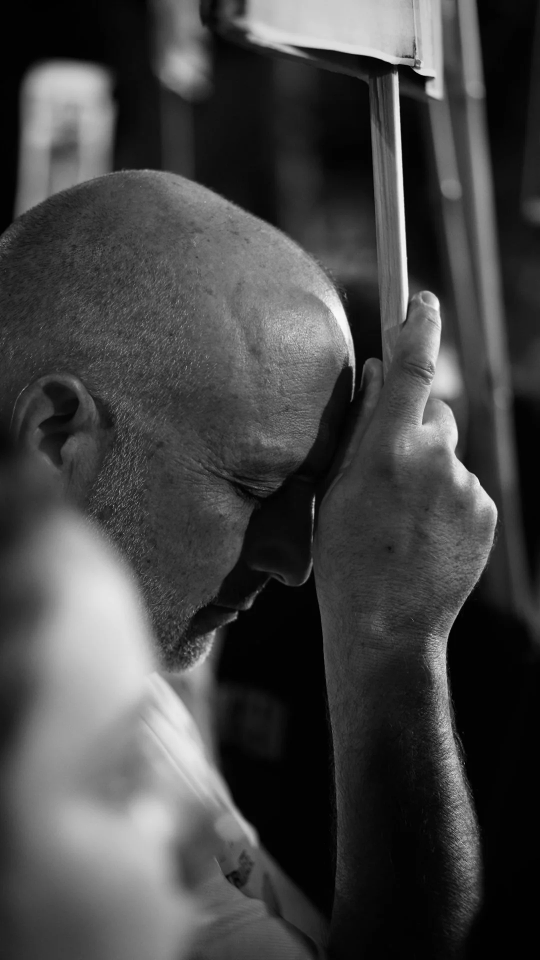 A man with a shaved head and a beard is resting his forehead on his hand while sitting on a bus, holding onto a bus handrail.