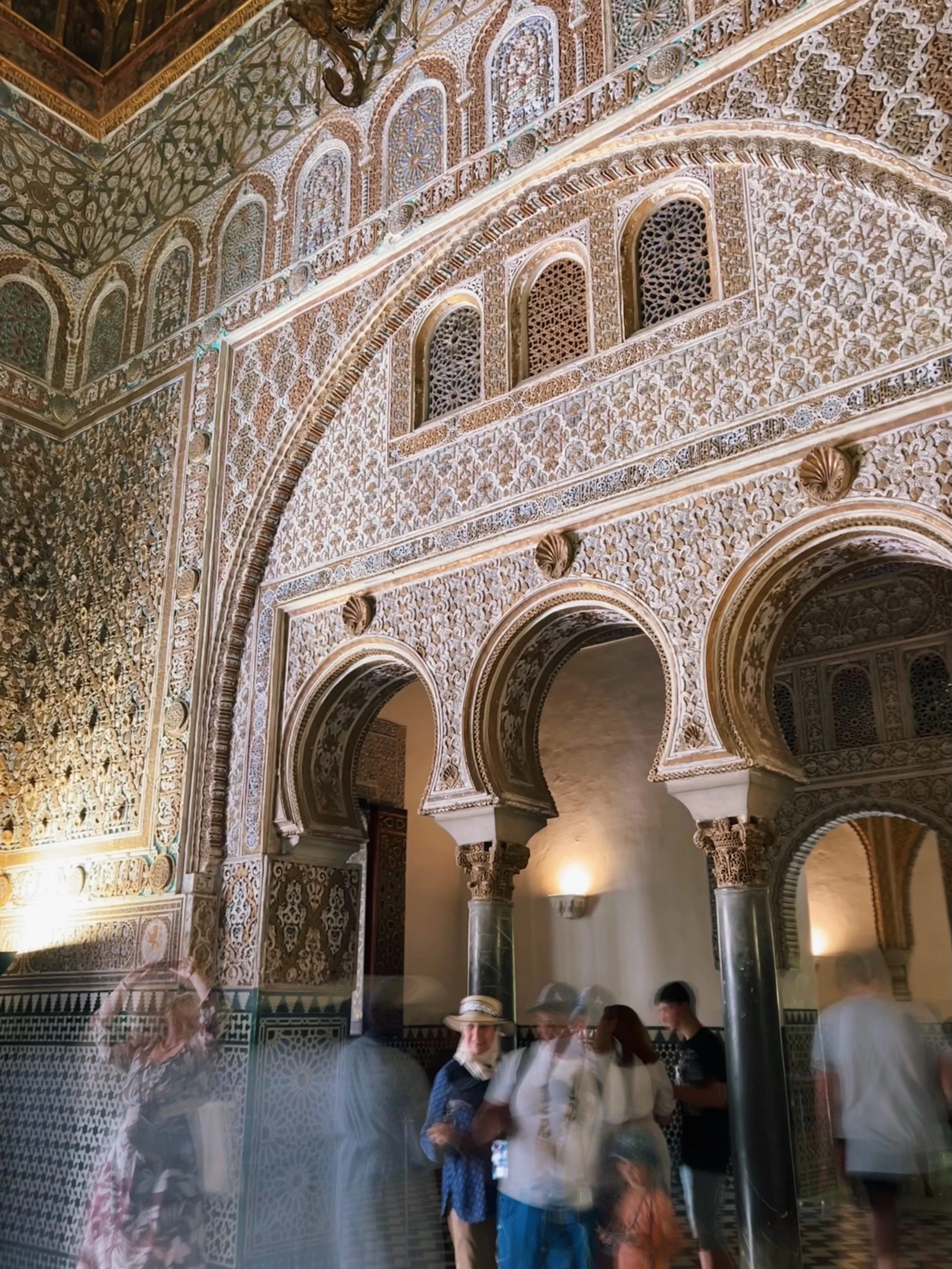 Intricate Islamic architectural interior with ornate arches and detailed decorative patterns, and a group of people, some blurred, standing inside.