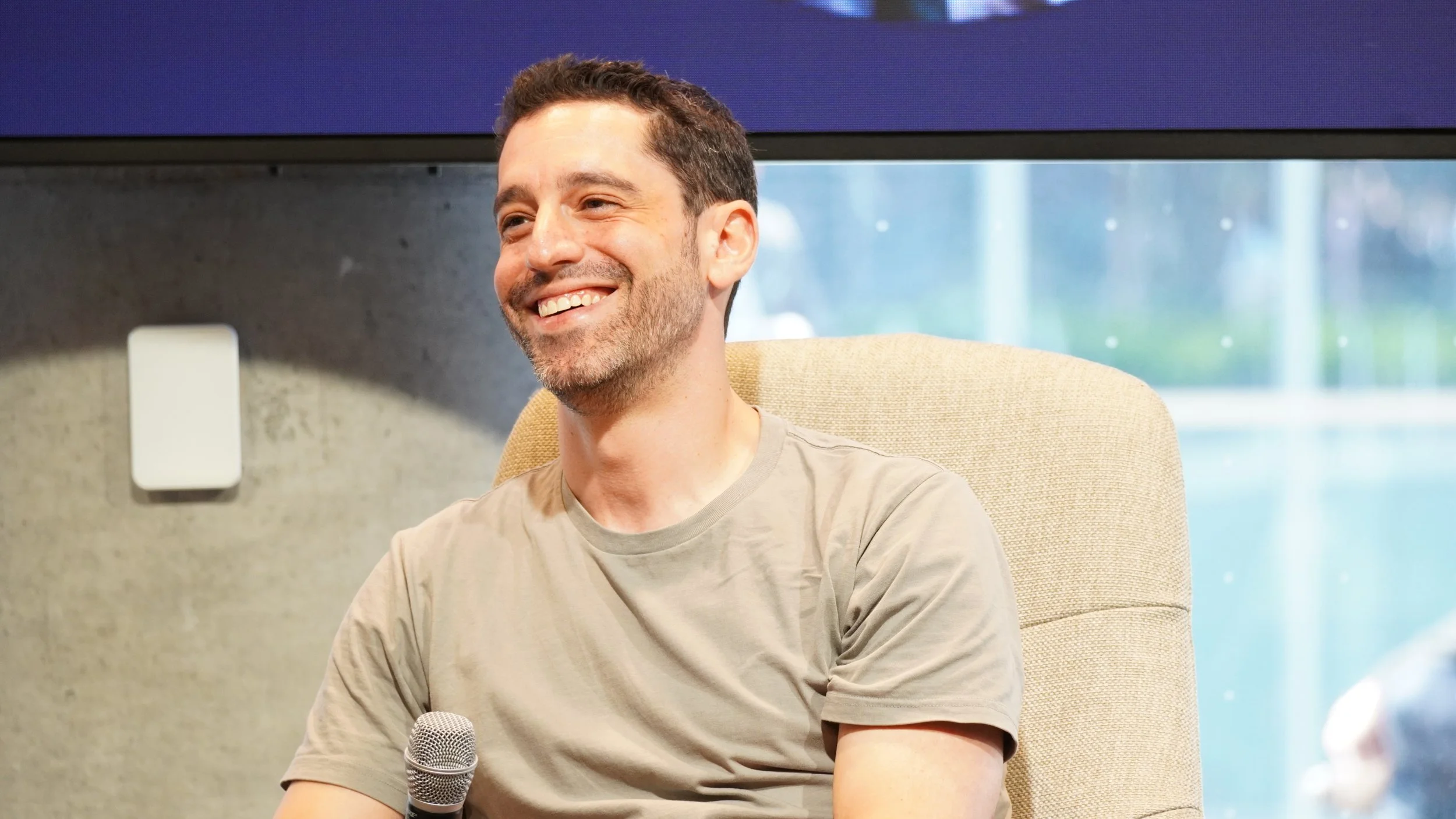A smiling man with a beard and short brown hair, sitting in a beige chair, holding a microphone, in a setting with a gray wall and a window with a blurred view outside.