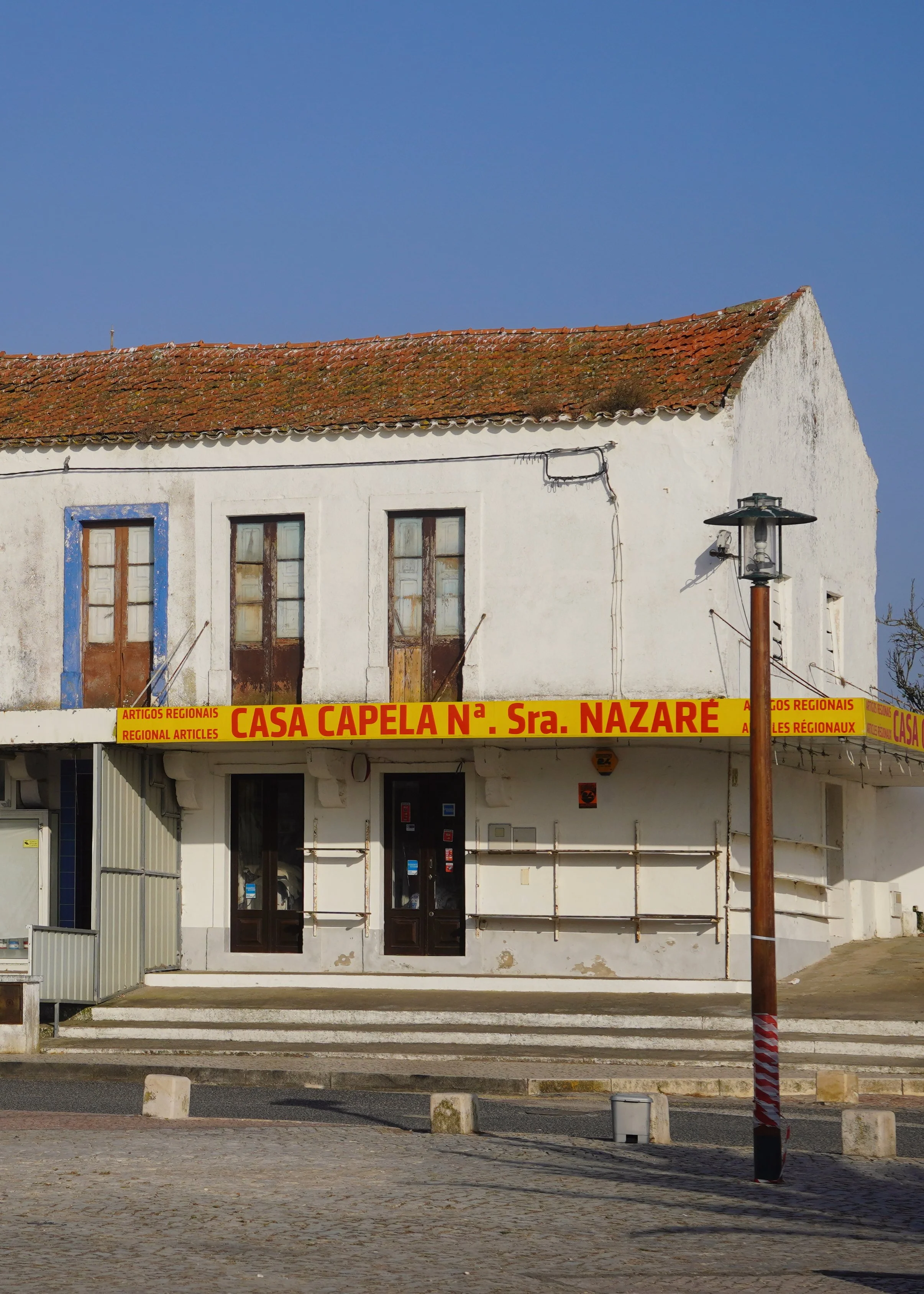 A white building with a red-tiled roof, brown doors, and windows. Yellow sign with red text reading 'Casa Capela N. Sra. Nazaré' and 'Artigos Regionais'.