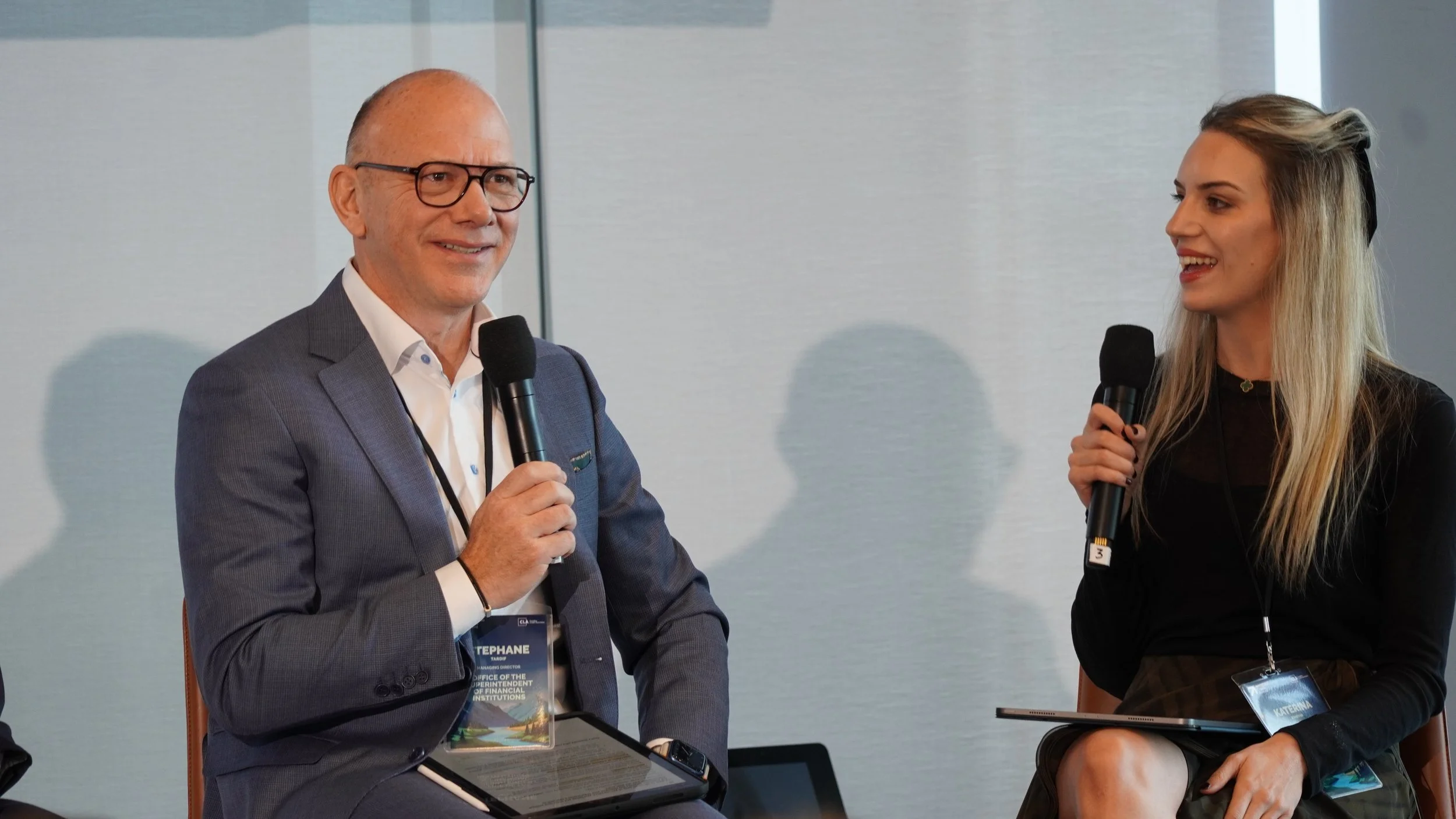 A man and woman sitting on stage with microphones, engaging in a conversation at a conference or panel discussion.