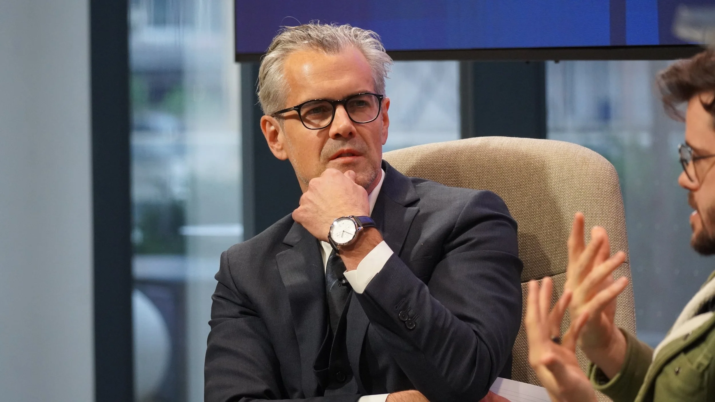 A man with gray hair, glasses, dressed in a business suit, sits with his hand on his chin, listening intently during a discussion. Two people are partially visible on his right, one of whom is gesturing with hands.