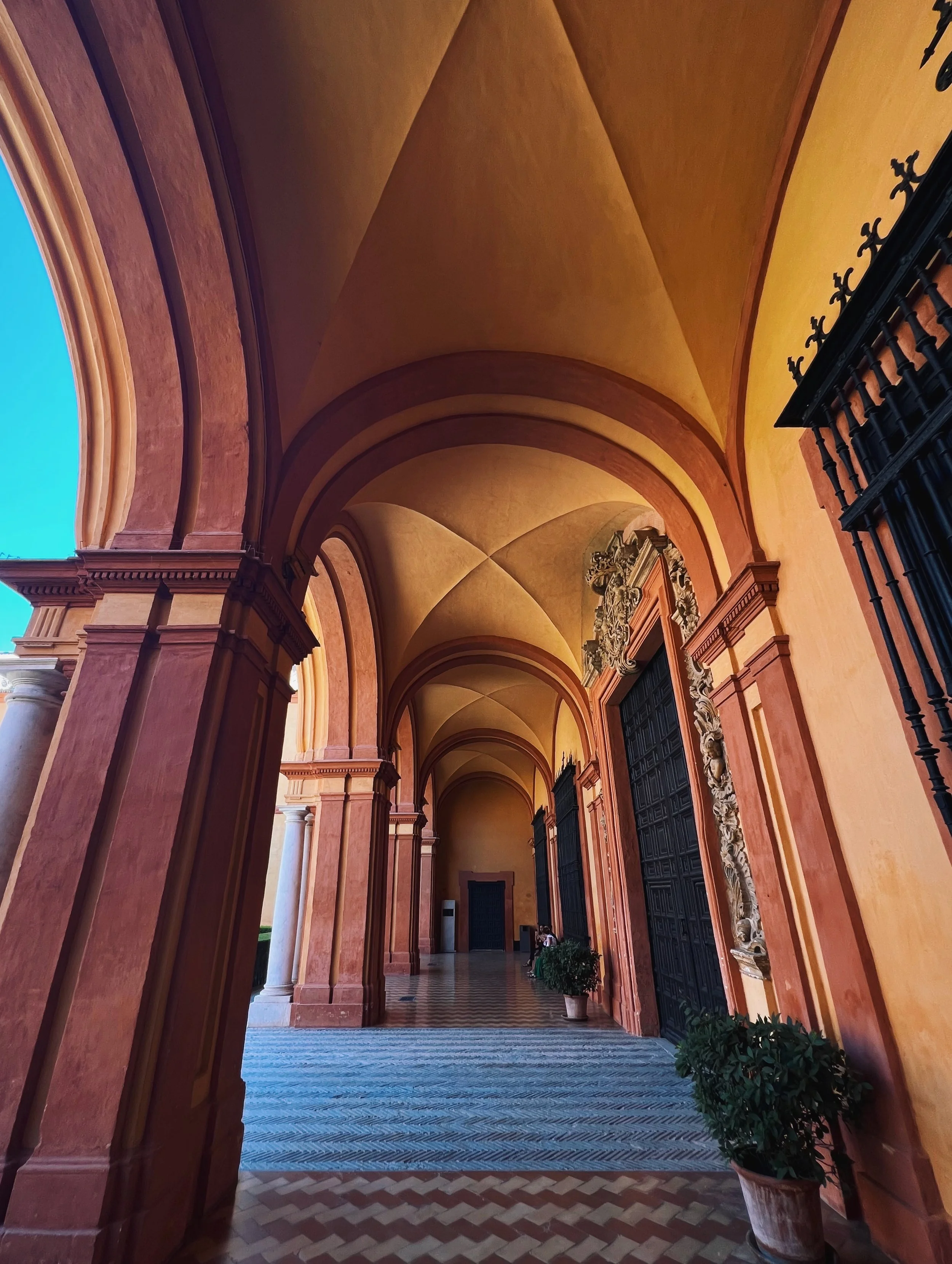 View of a covered porch with arched ceilings, ornate details, potted plants, and large black doors at the end.