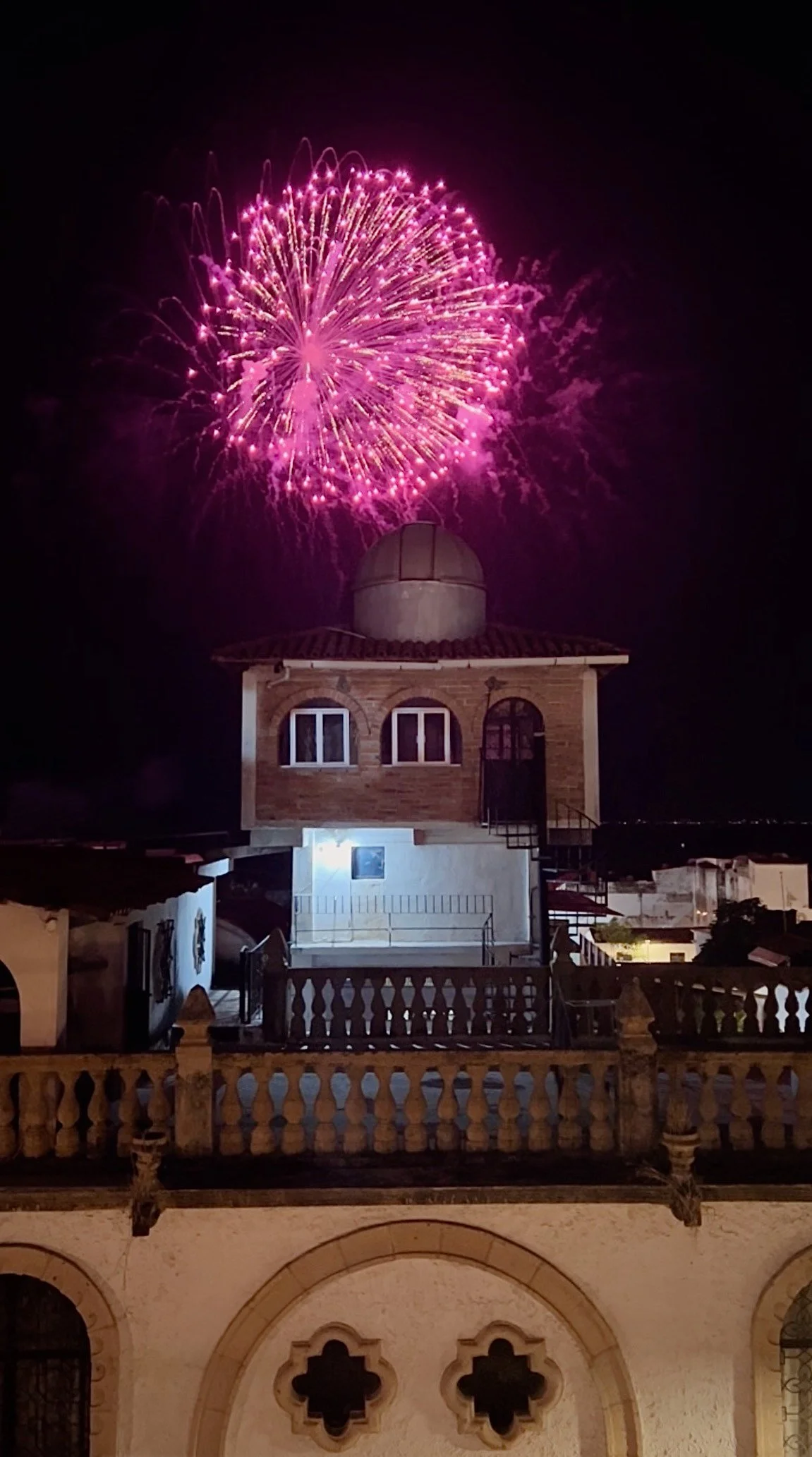 Pink fireworks exploding above an observatory building on a dark night. The building has a dome on top and brick walls with arched windows.
