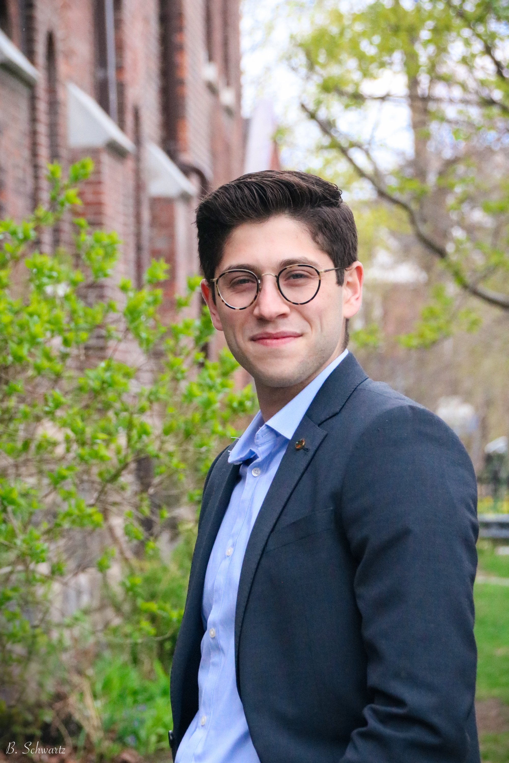 Young man in a blue shirt and blazer standing outdoors near a brick building and green trees, smiling at the camera.