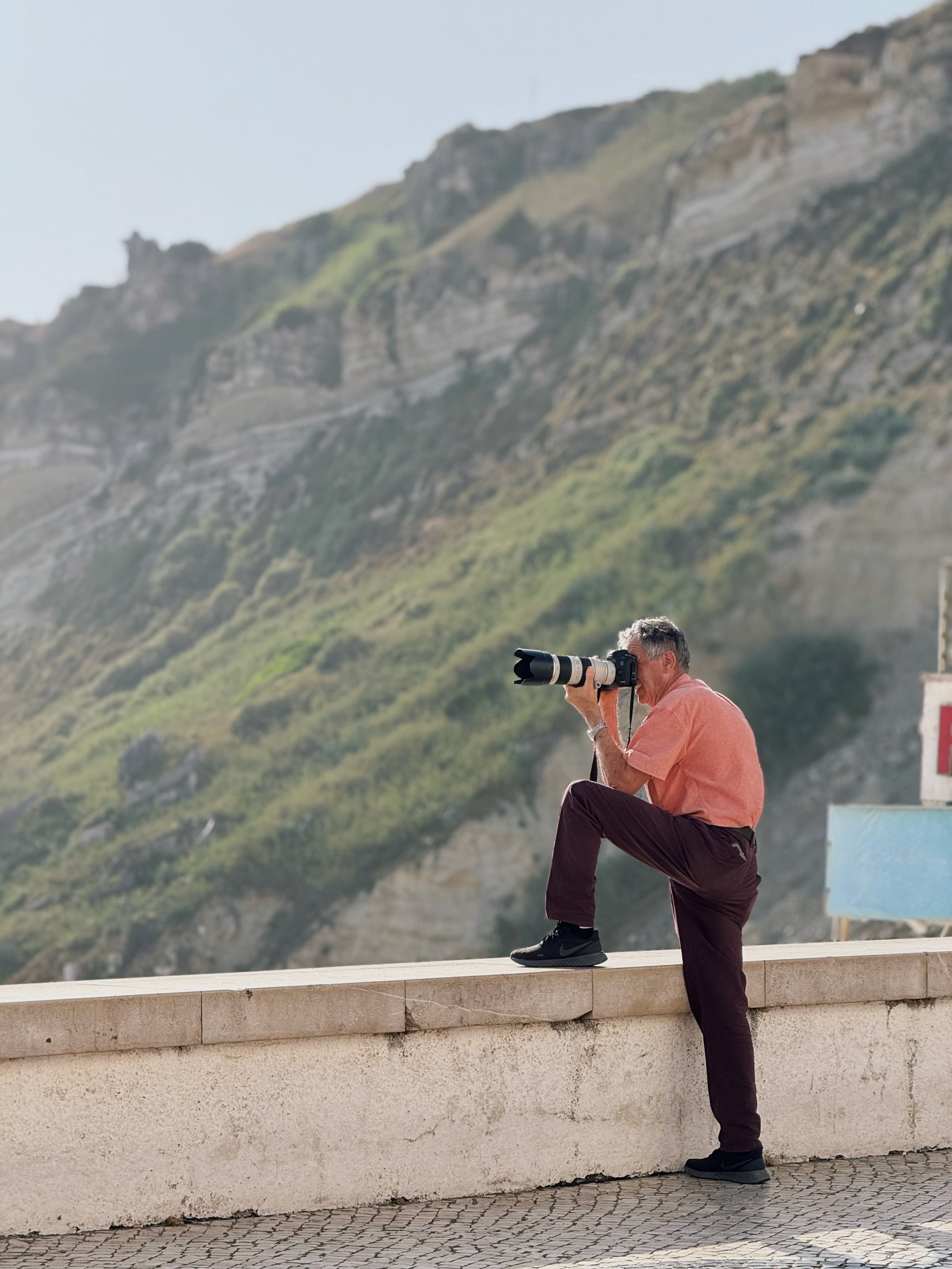 A man wearing a pink shirt and dark pants is standing on a beige stone ledge and taking photos with a large telephoto lens camera, with a mountain hillside in the background.