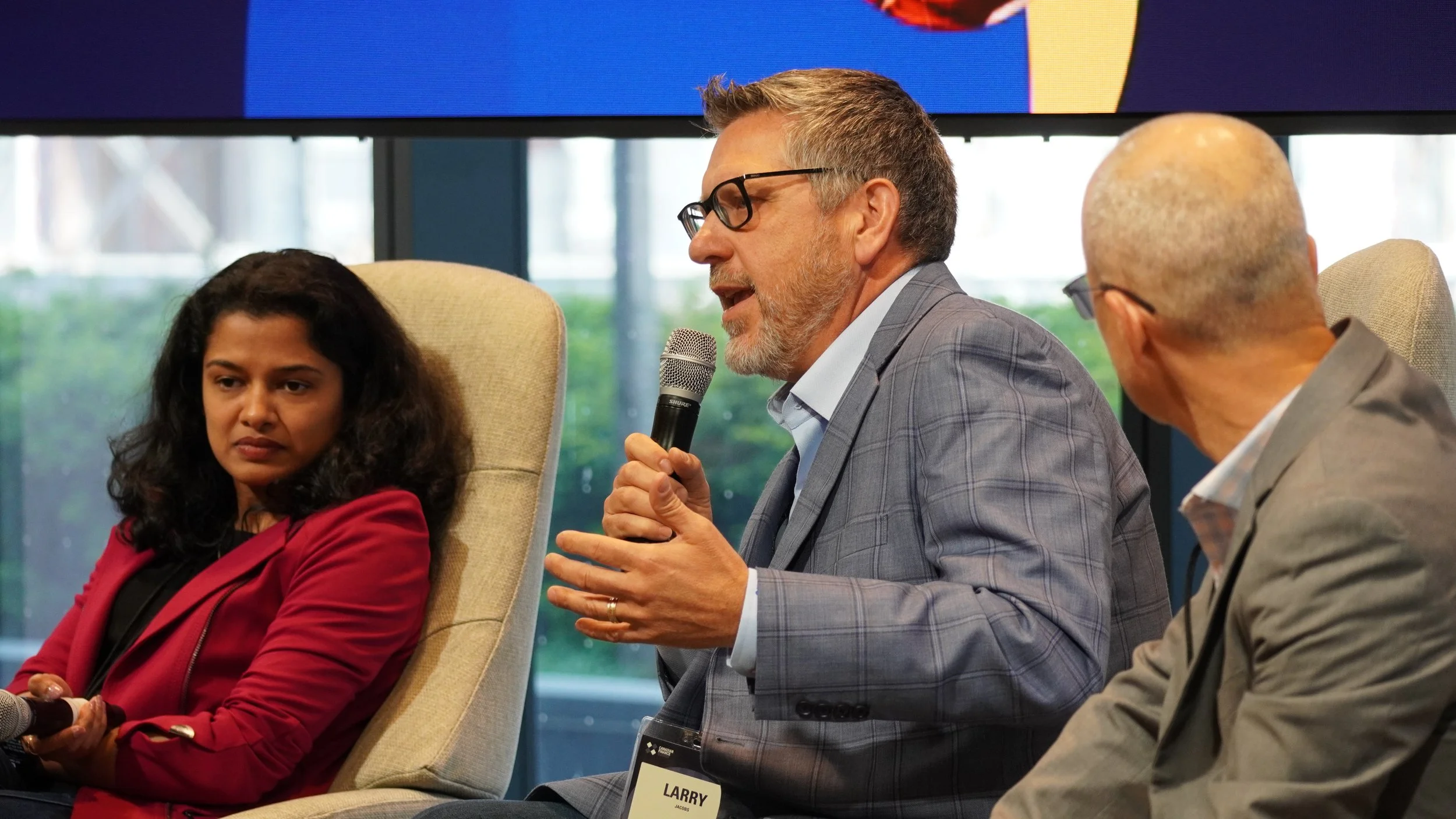 Three professionals seated on a panel during a discussion or conference. The man in the middle, wearing glasses and a checkered suit, is speaking into a microphone. The woman on the left, dressed in a red blazer, appears to be listening thoughtfully.