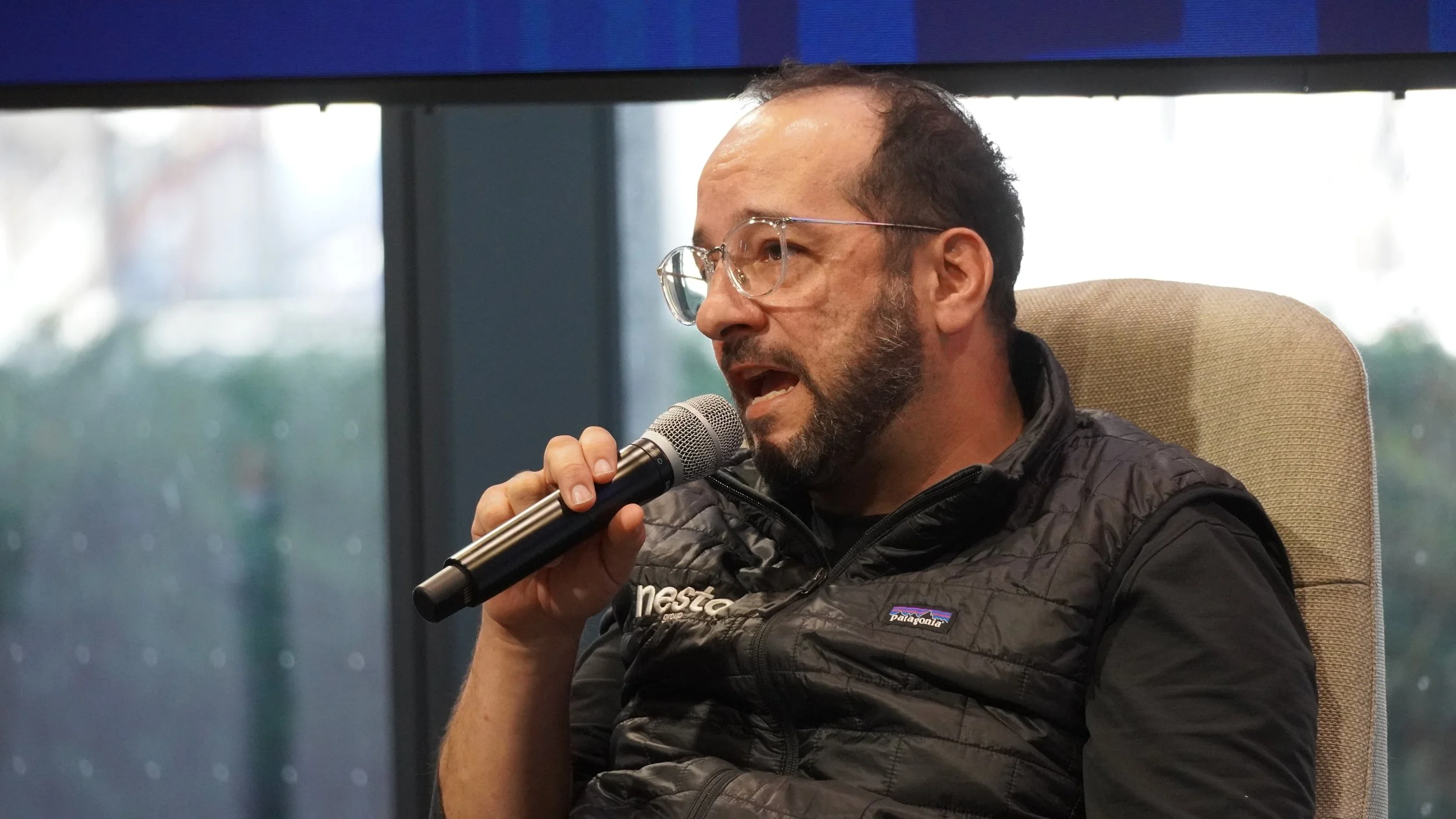 A man with glasses and a beard is speaking into a microphone while sitting on a beige chair. He is wearing a black Patagonia vest and a black shirt.