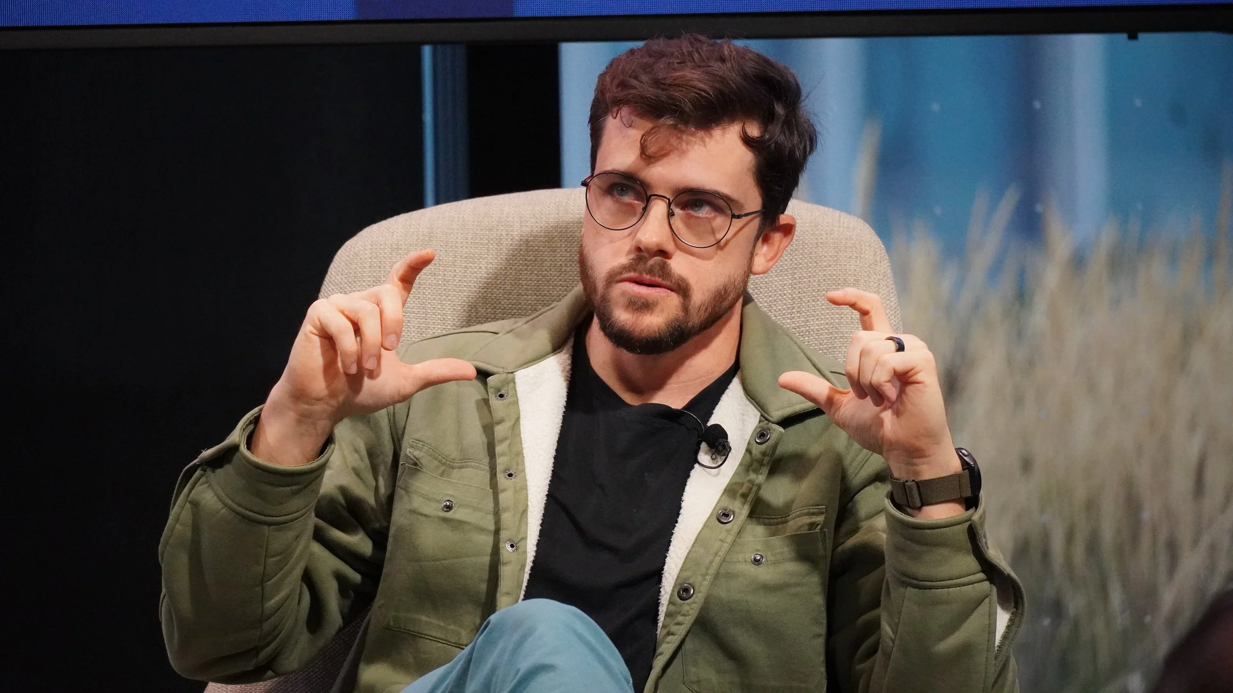 A man with glasses, a beard, and short brown hair, wearing a green jacket and a black shirt, is sitting in a chair, gesturing with both hands, in an indoor setting.