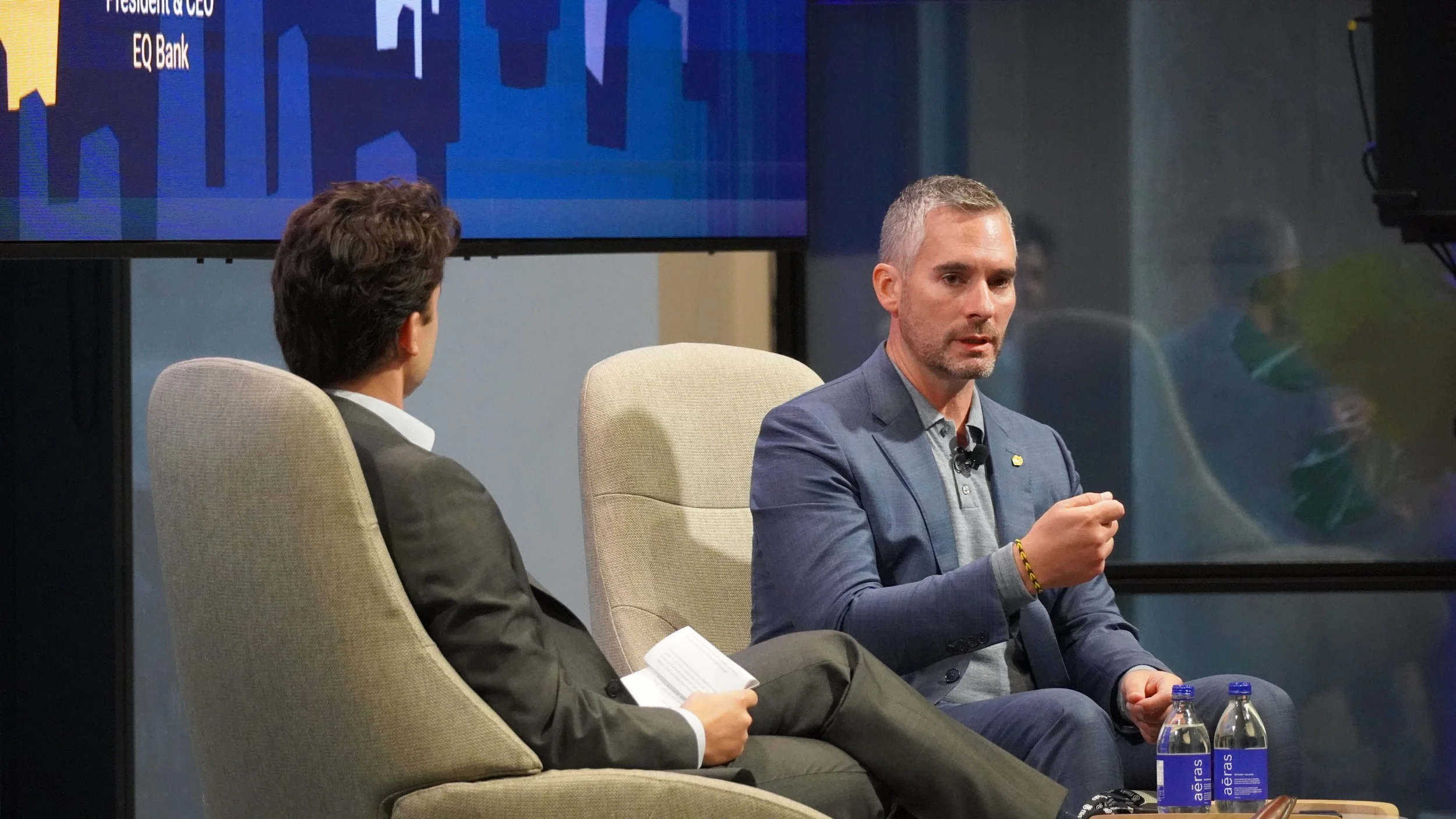 Two men sitting on beige chairs engaged in a conversation or interview on a stage or panel. The man on the right is speaking, gesturing with his right hand. The background shows a large screen with part of a logo or graphic, and two water bottles are