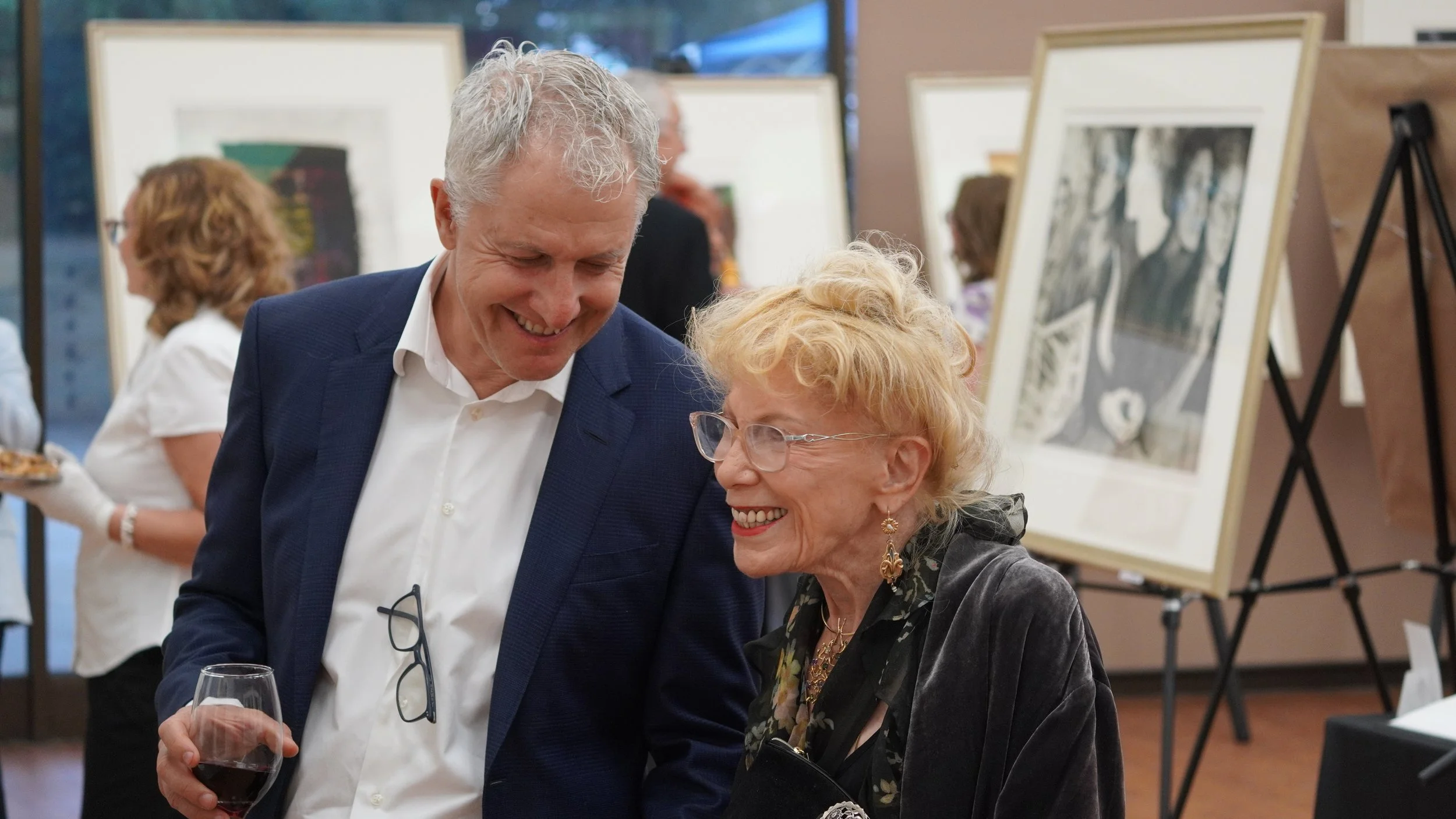 A man and woman smiling and talking at an art gallery event. The man holds a glass of red wine and wears a navy blazer, white shirt, and glasses hanging from his shirt. The woman wears glasses, earrings, a dark jacket, and a scarf. In the background,