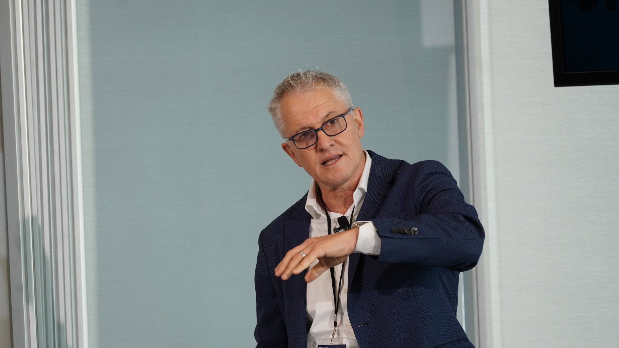 A man with gray hair and glasses wearing a dark blue blazer and white shirt, talking and gesturing with his hand in an office setting.