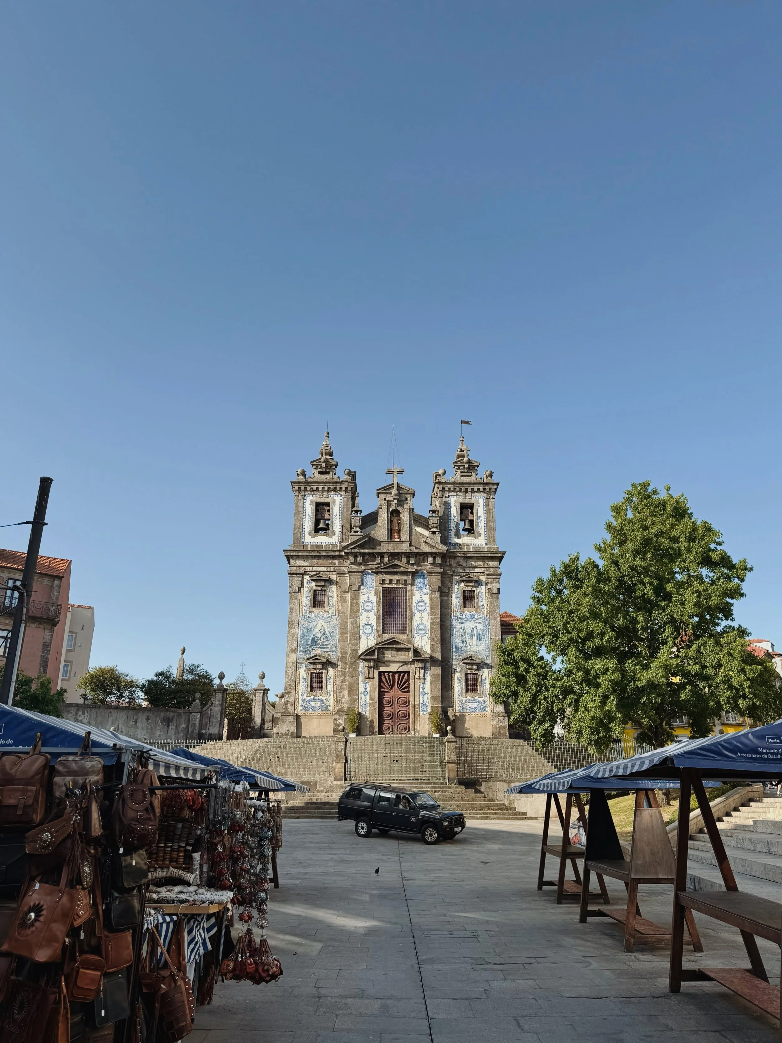 A historic church with ornate architecture, towers, and a cross at the top, situated on a staircase in a plaza with street stalls and a tree on the right, under a clear blue sky.
