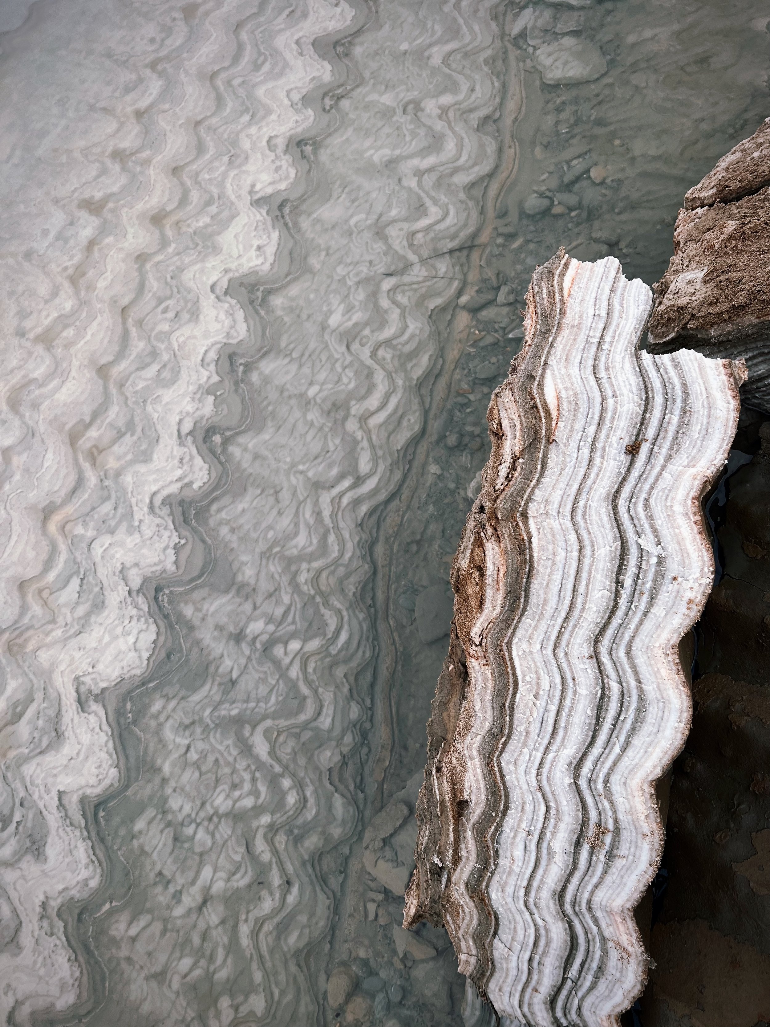 Close-up of layered rock formations with wavy, striped patterns in shades of white, gray, and brown, next to water with more layered rock patterns visible.