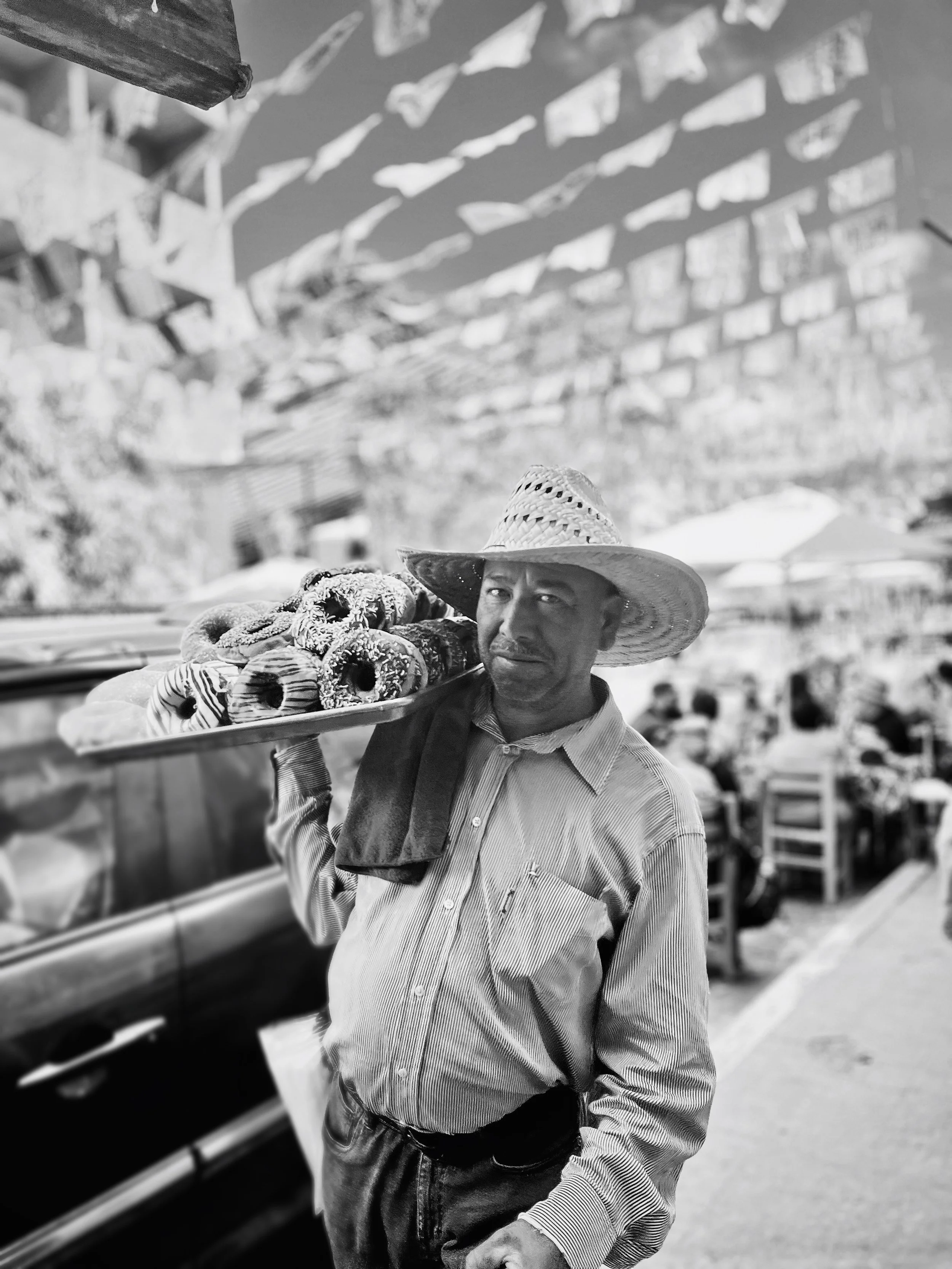 A man wearing a large straw hat and striped shirt carrying a tray of donuts at an outdoor market or festival.